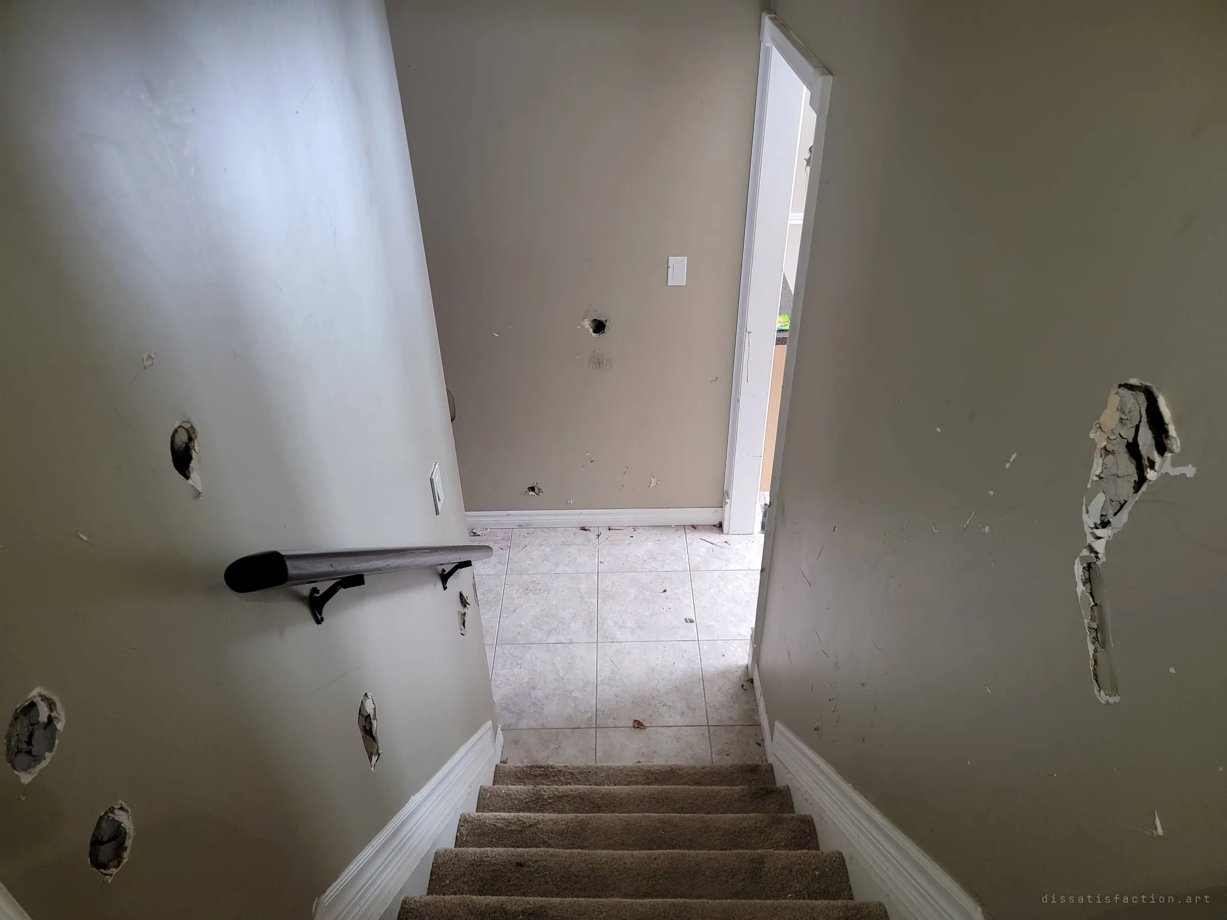 Looking down a carpeted staircase between walls pocked with punched holes toward a tiled landing and an open doorway. The damage follows the whole way down.