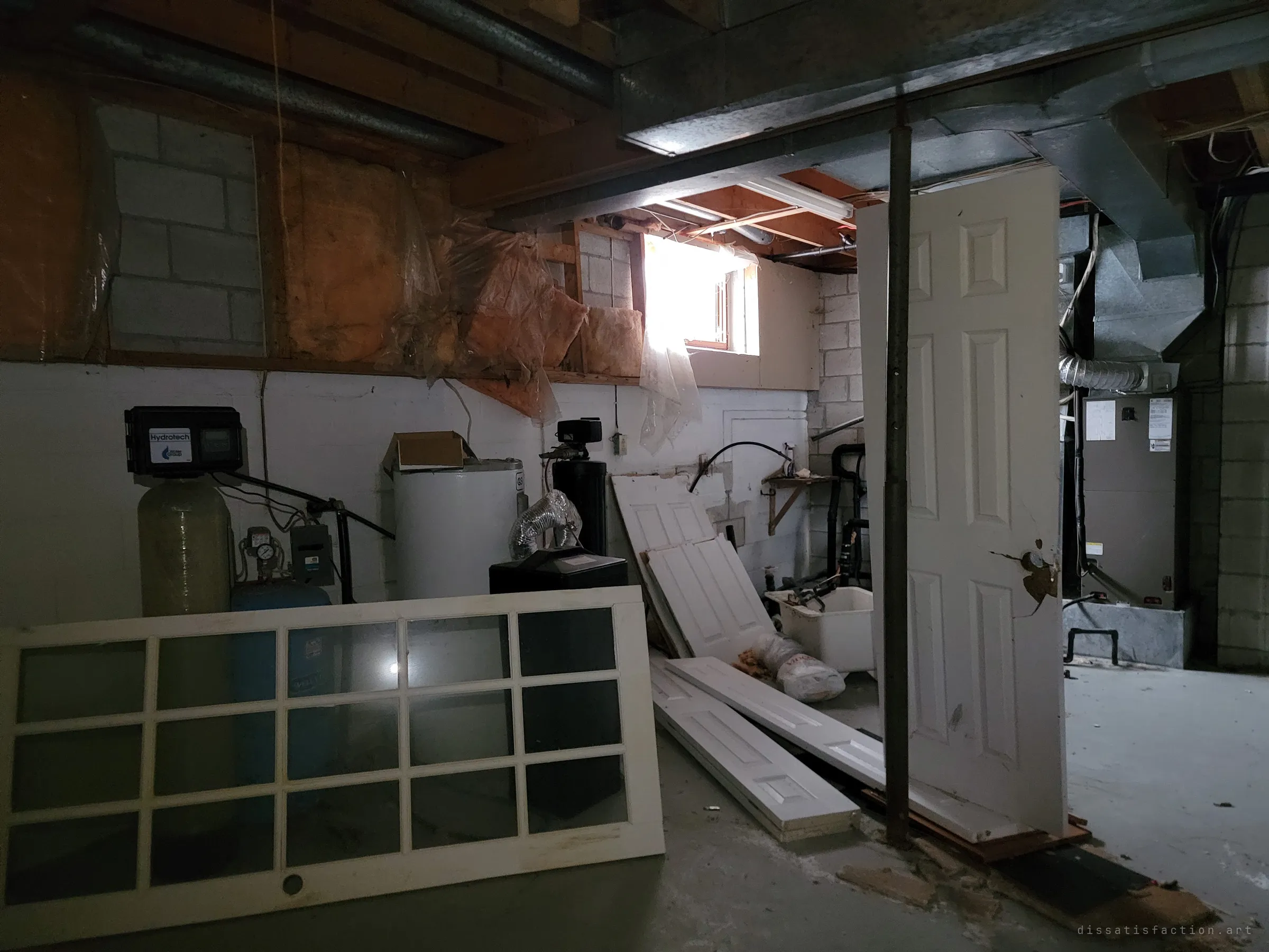 An unfinished basement with a water softener, water heater and furnace under exposed ductwork and insulation, loose doors and a glazed French door stacked against the utilities. Daylight from a small basement window cuts through the gloom.