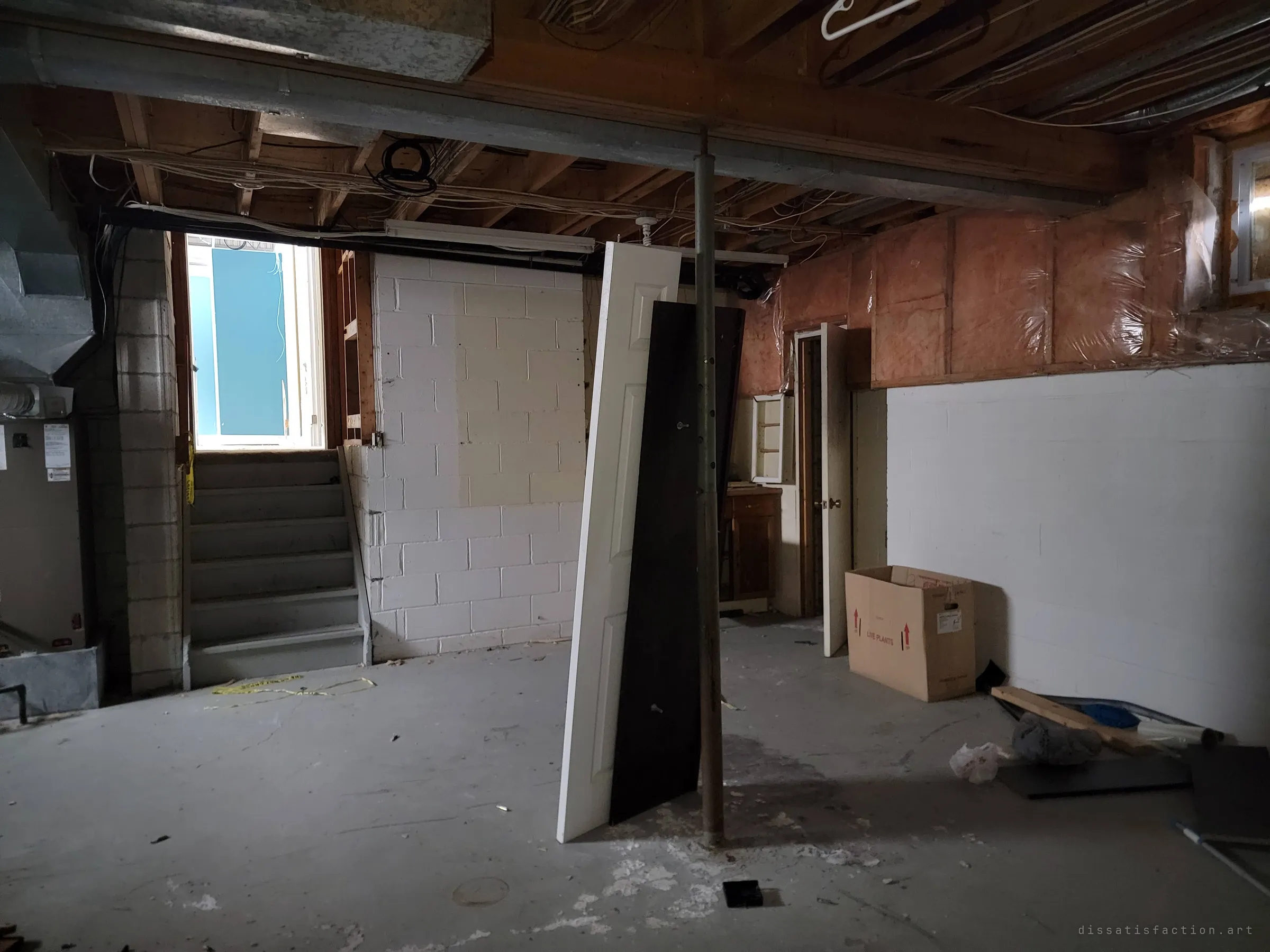 The open basement floor with cinder-block walls, a steel support post propping up two leaning doors, a cardboard box and scraps of yellow caution tape near the stairs up to a bright window well. Empty and stripped, the space feels abandoned rather than lived in.