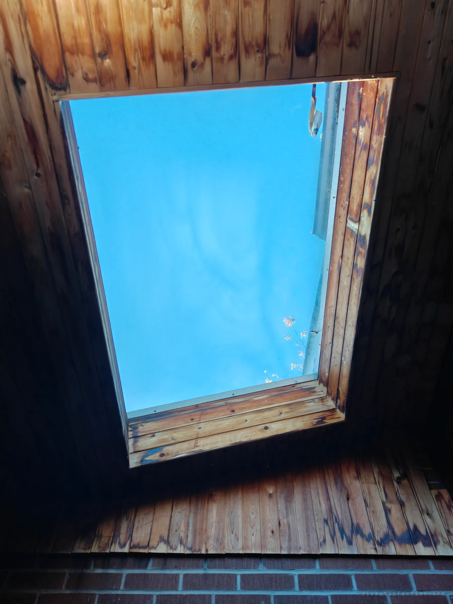 Looking up through a skylight set in scorched wood-panelled ceiling boards, with bright blue sky and a few bare twigs in the corner above a brick wall.