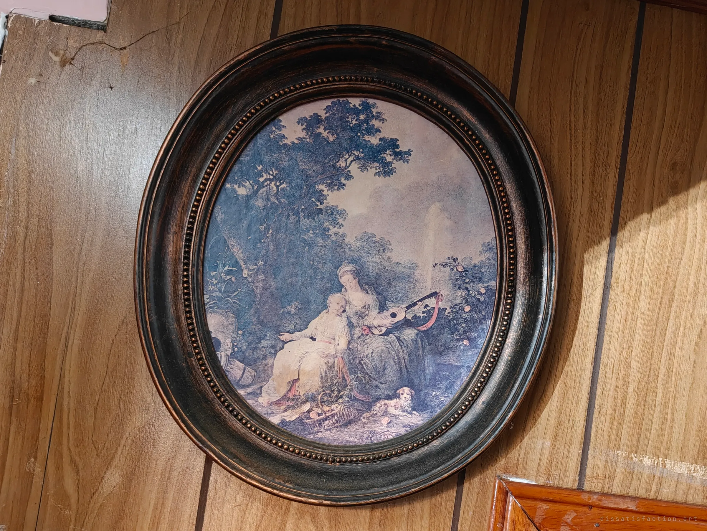 A dark oval frame with a beaded edge hangs on wood-panelled wall, holding a faded print of two women with a lute and a small dog in a garden.