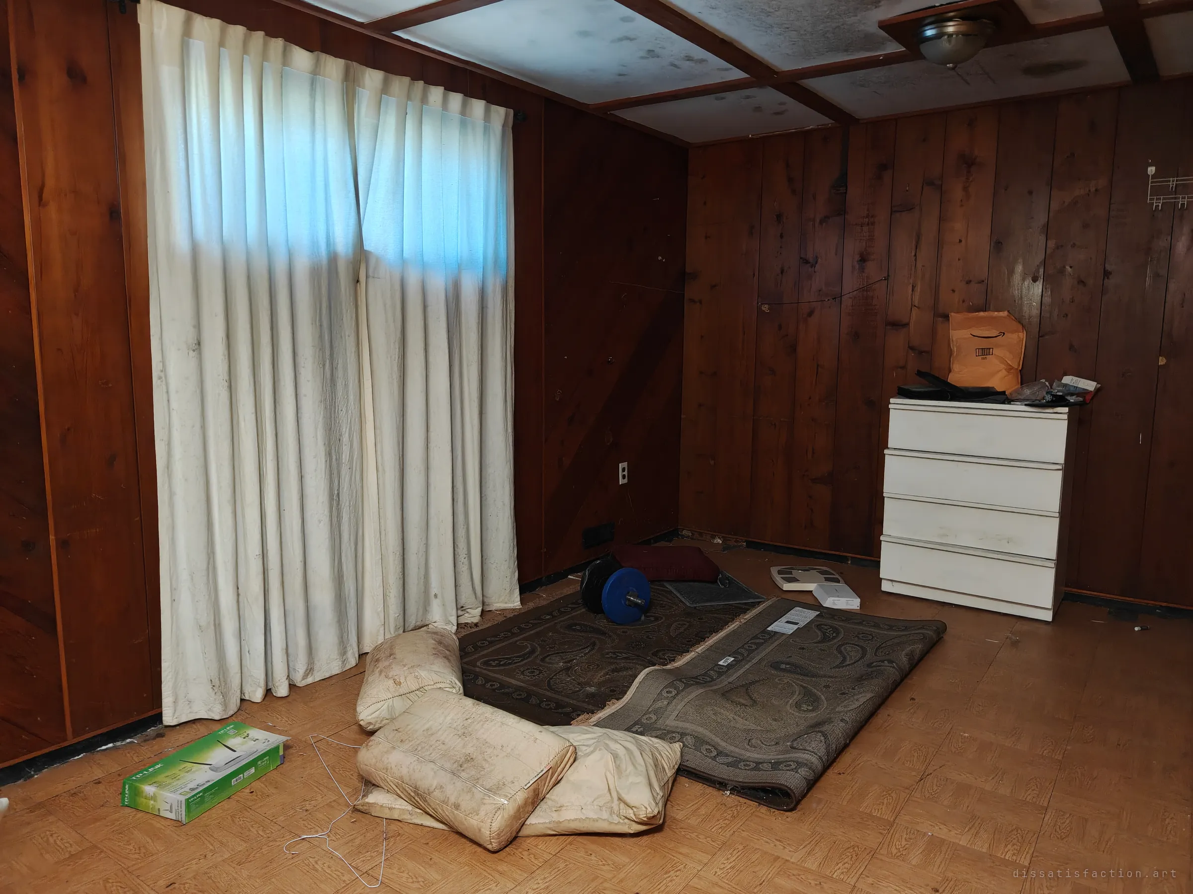 A bare wood-panelled room with grimy white curtains over a window, a worn rug, stained pillows, a barbell plate, a router box and a white chest of drawers.