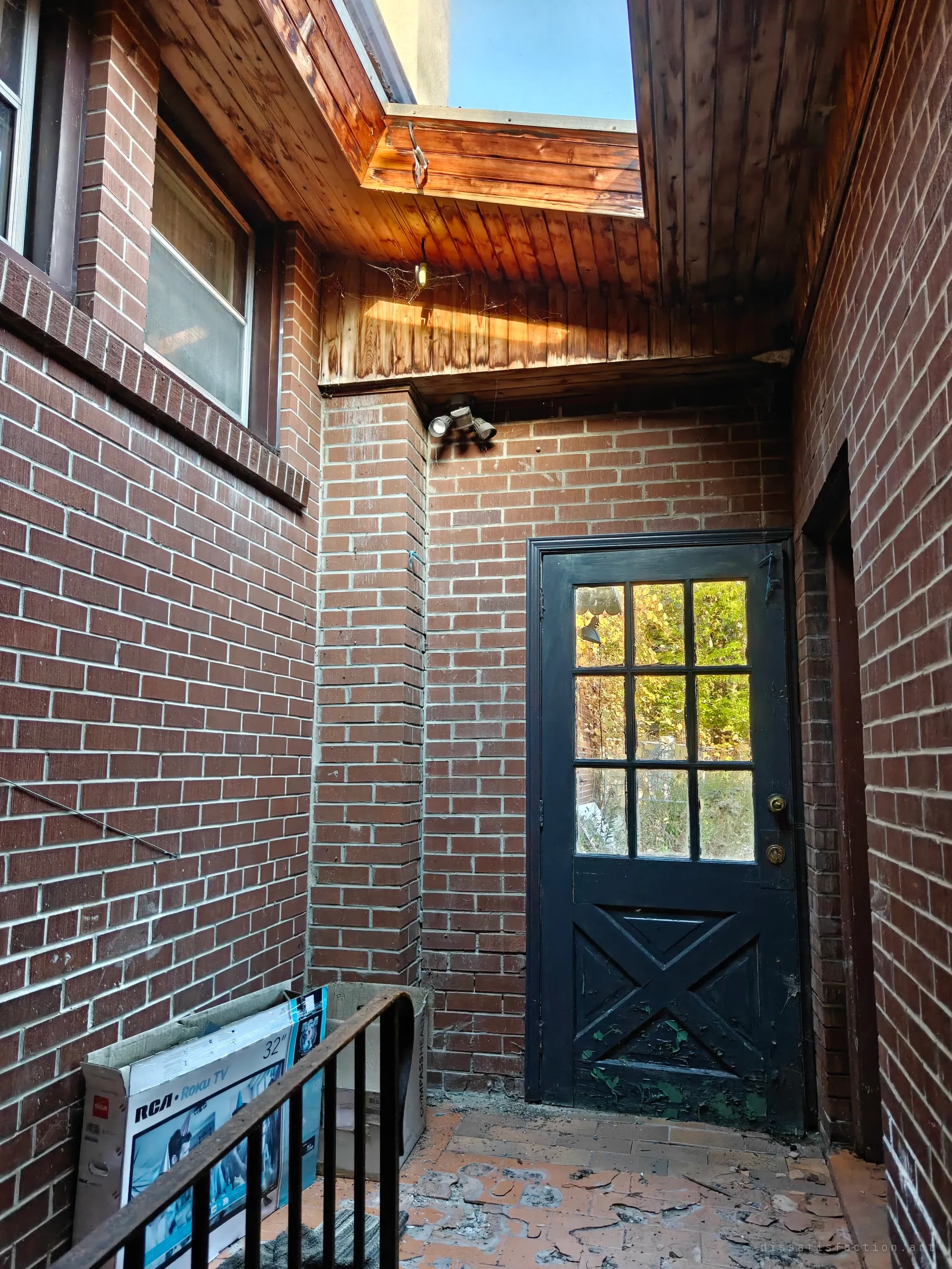 A small brick courtyard with a black glazed door, a flood light under scorched timber soffit and a gap to the sky, an RCA TV box and an iron railing in the foreground, and flaking tile underfoot.