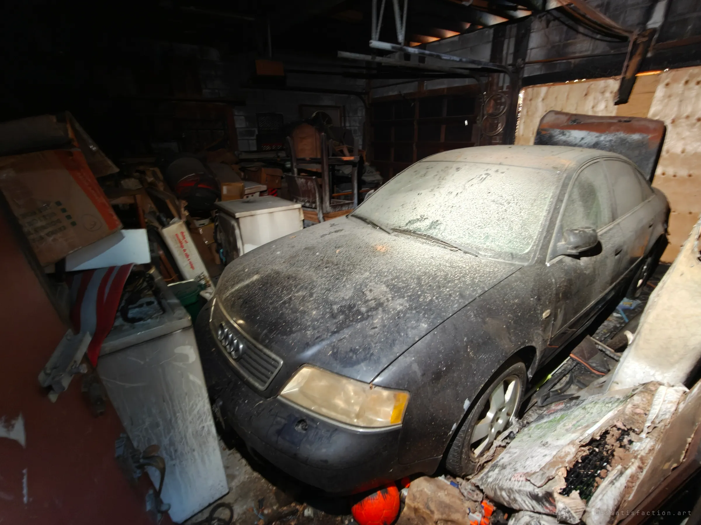 A dust-covered dark Audi sedan parked in a fire-damaged garage, hemmed in by scorched furniture, boxes and appliances, with a charred board leaning against it.