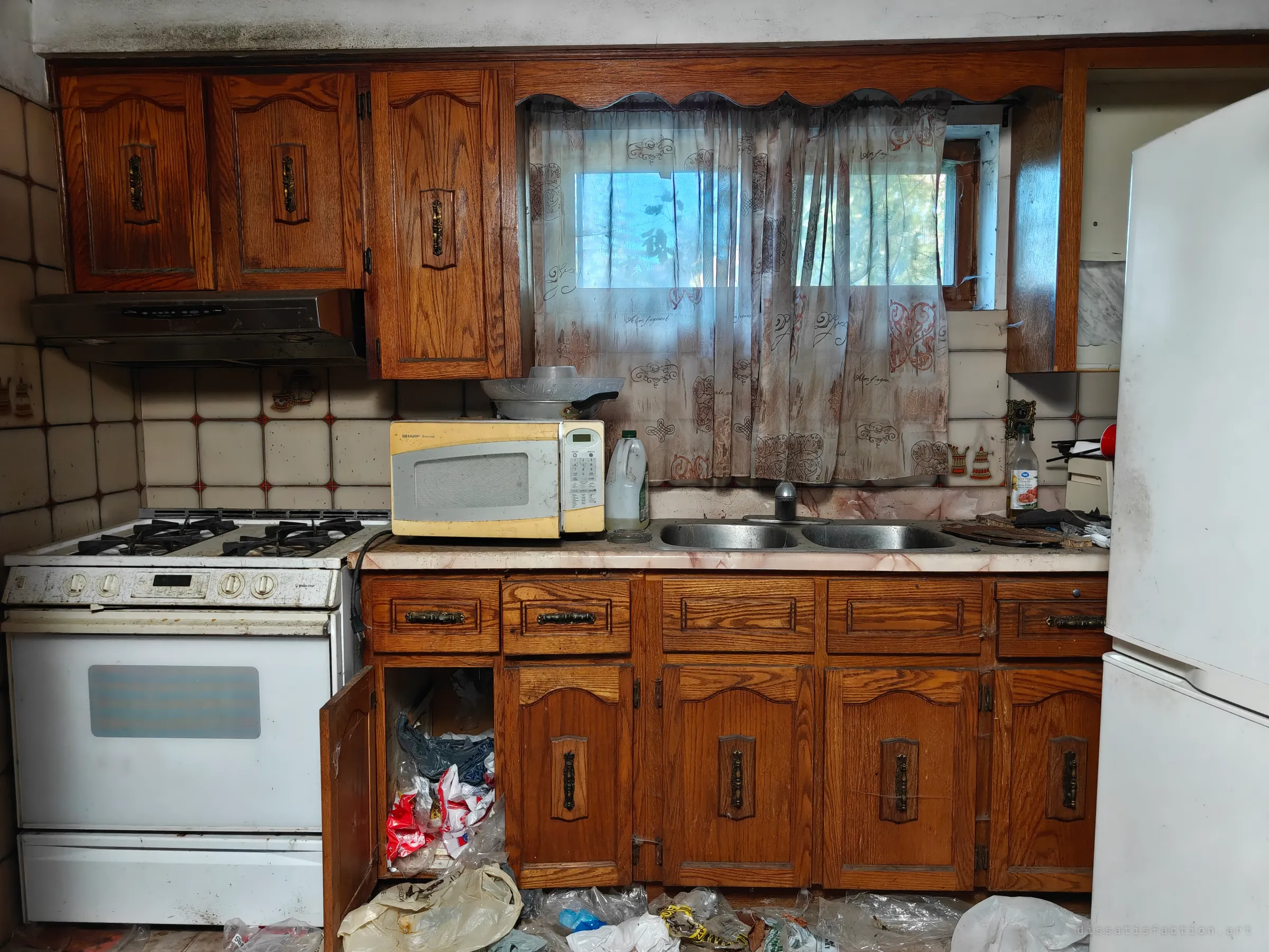 A dated oak kitchen with a sheer curtain over a small window, a gas stove, a microwave and open cabinet doors spilling plastic bags onto the floor.
