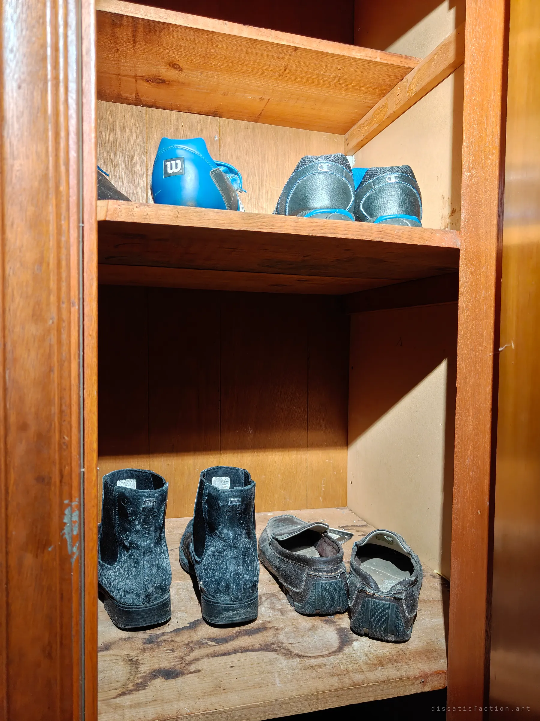 Two shelves of a wooden cupboard holding abandoned shoes: blue and grey trainers above, and two mould-speckled black boots and a pair of worn loafers below.