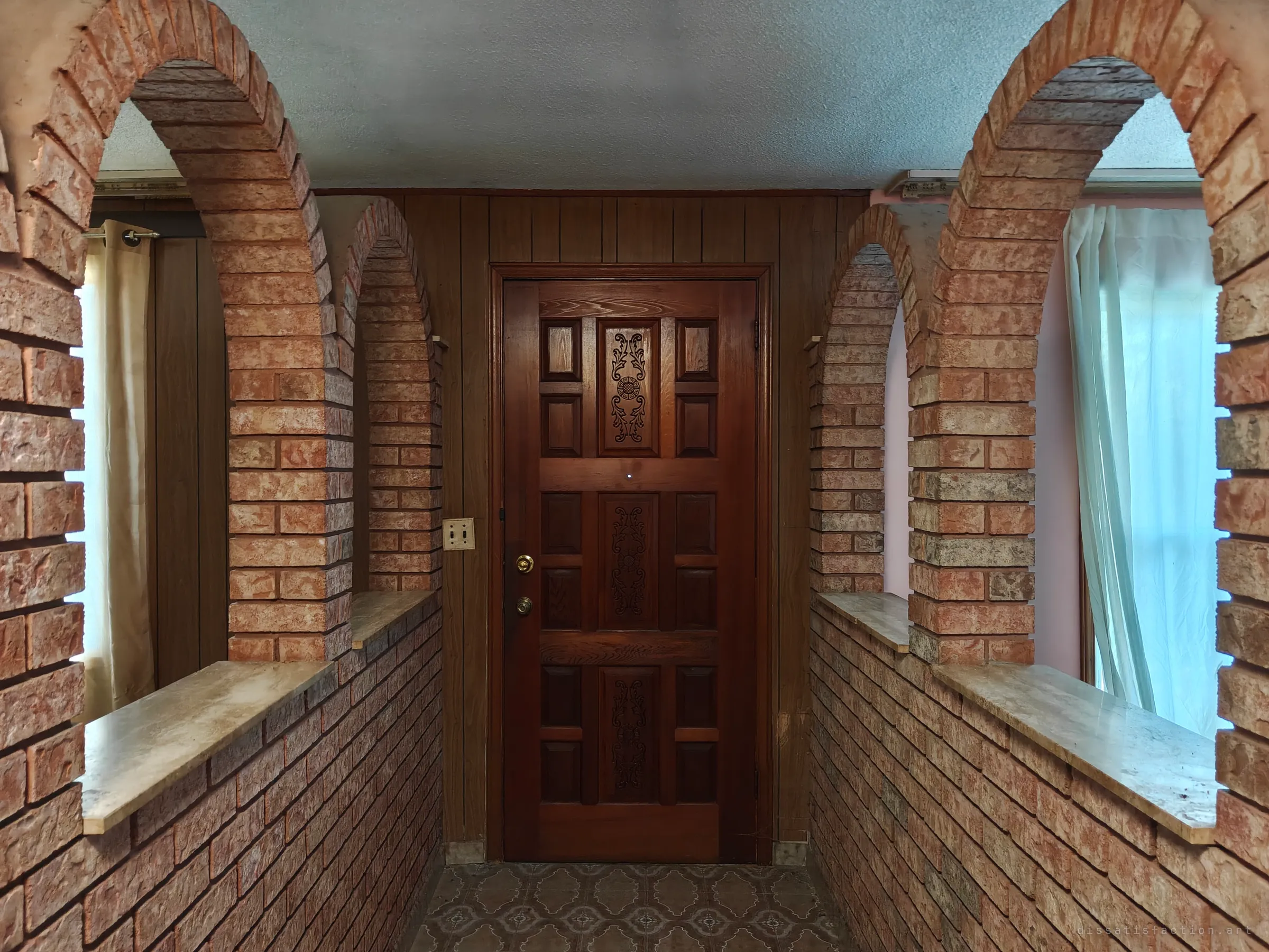 A narrow entry hall between two brick arcades with marble sills leads to a carved dark-wood front door, with curtained windows glowing at either side.