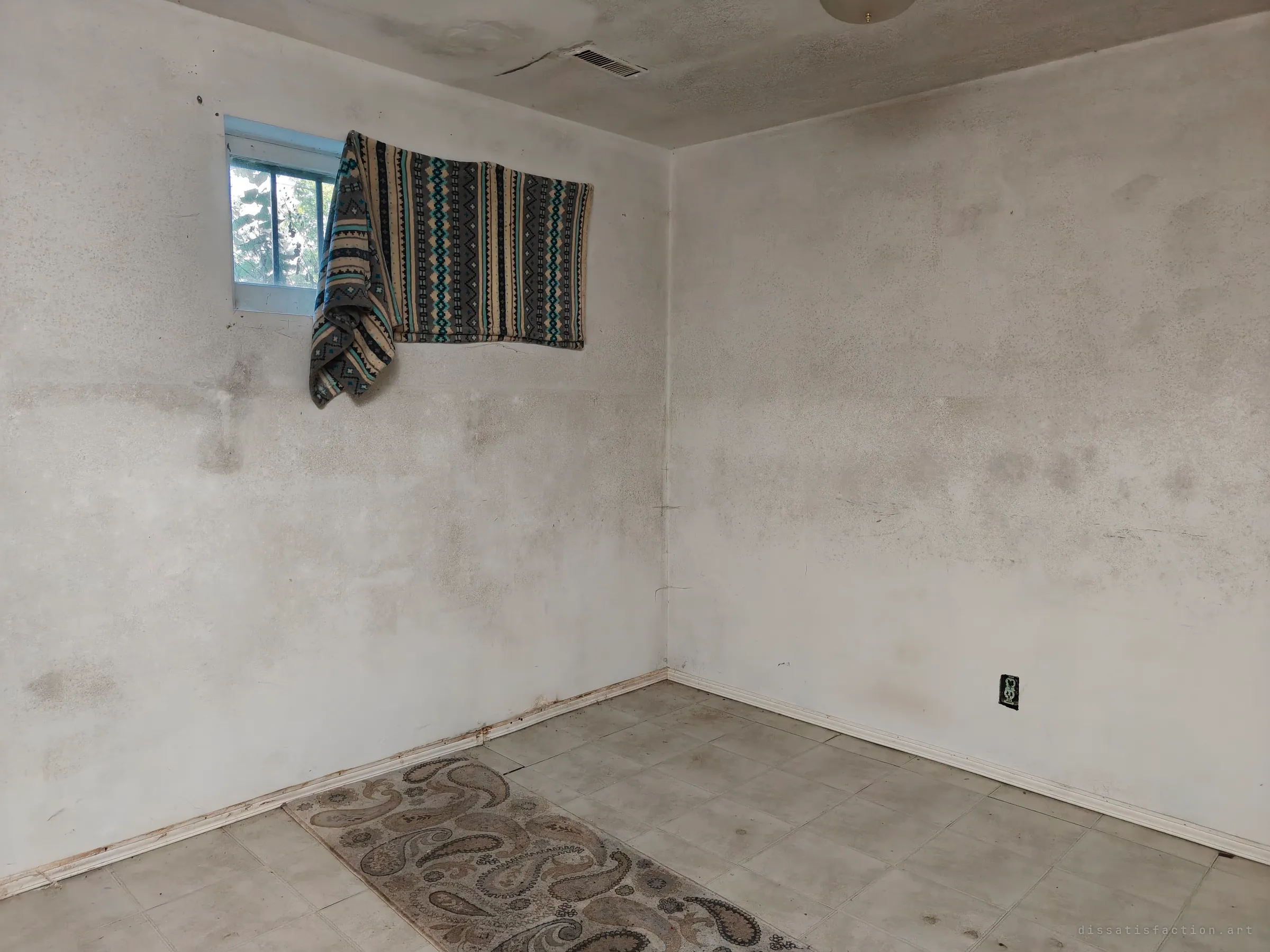 A bare, grimy basement room corner with a small high window half covered by a patterned blanket, and a worn paisley rug on pale tile floor.