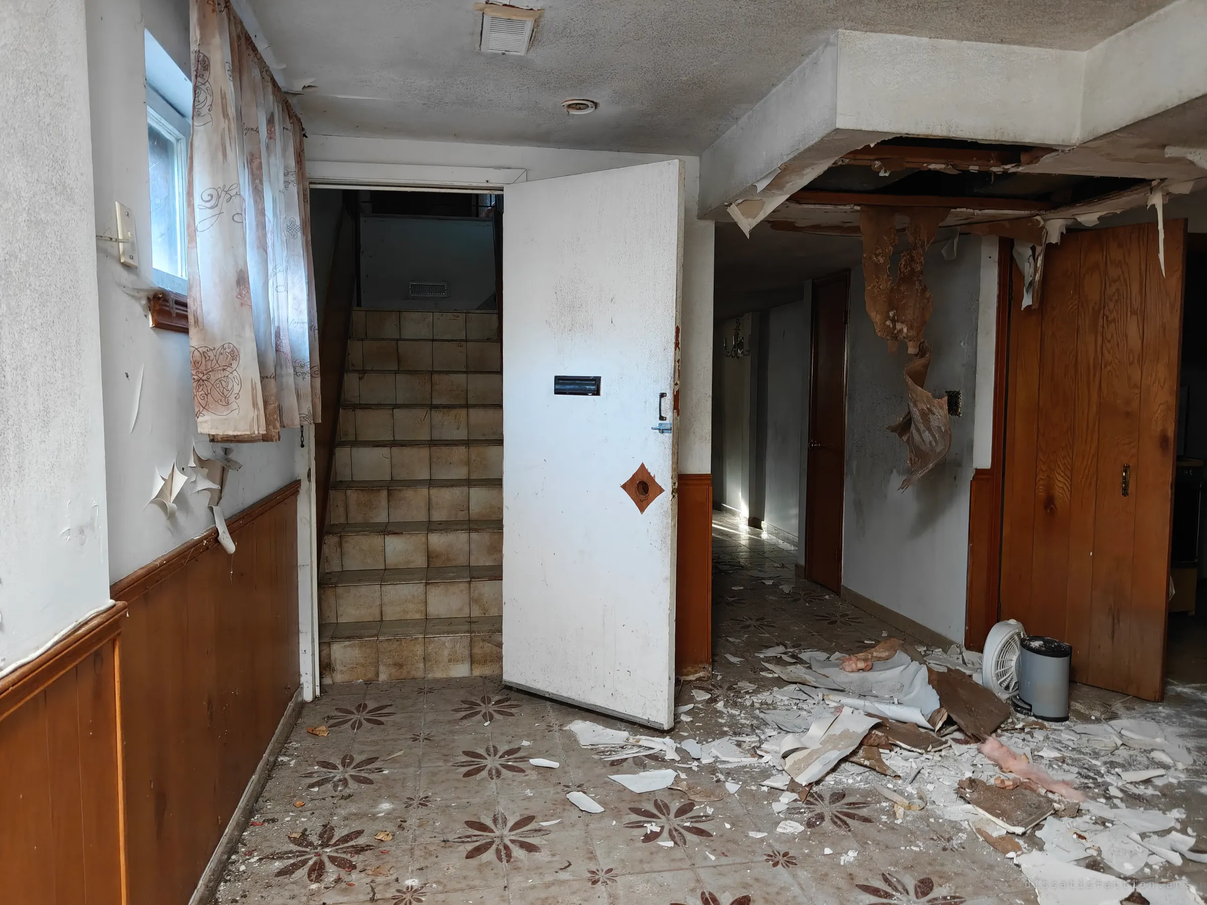 A basement hallway where plaster and ceiling paper have collapsed onto a patterned tile floor. A white door stands open beside a tiled staircase, with a long corridor running off to the right.