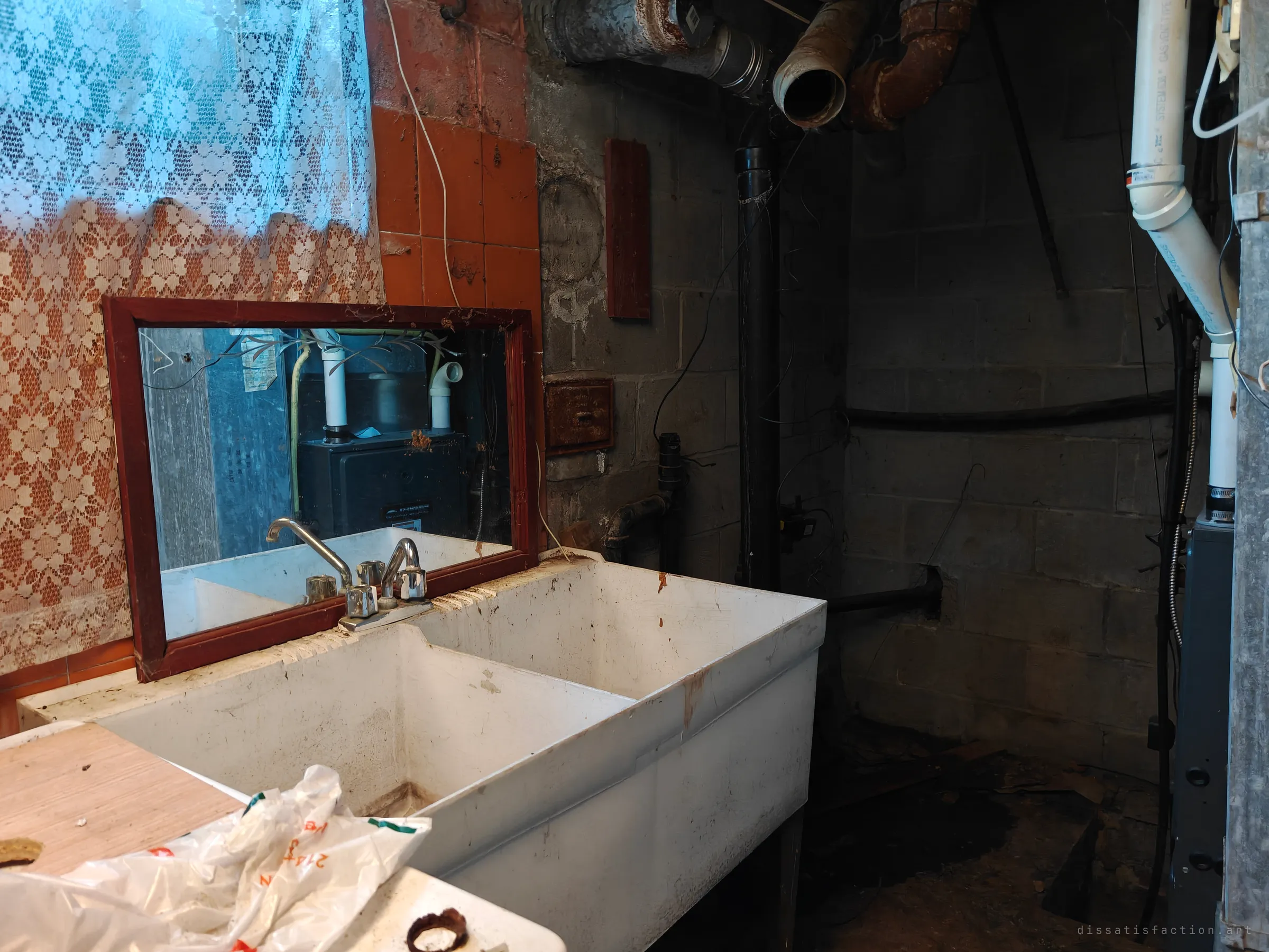 A grimy double laundry sink in a dark cinder-block basement corner. A lace curtain hangs at left, rusted pipes run overhead, and a mirror above the sink reflects a furnace.