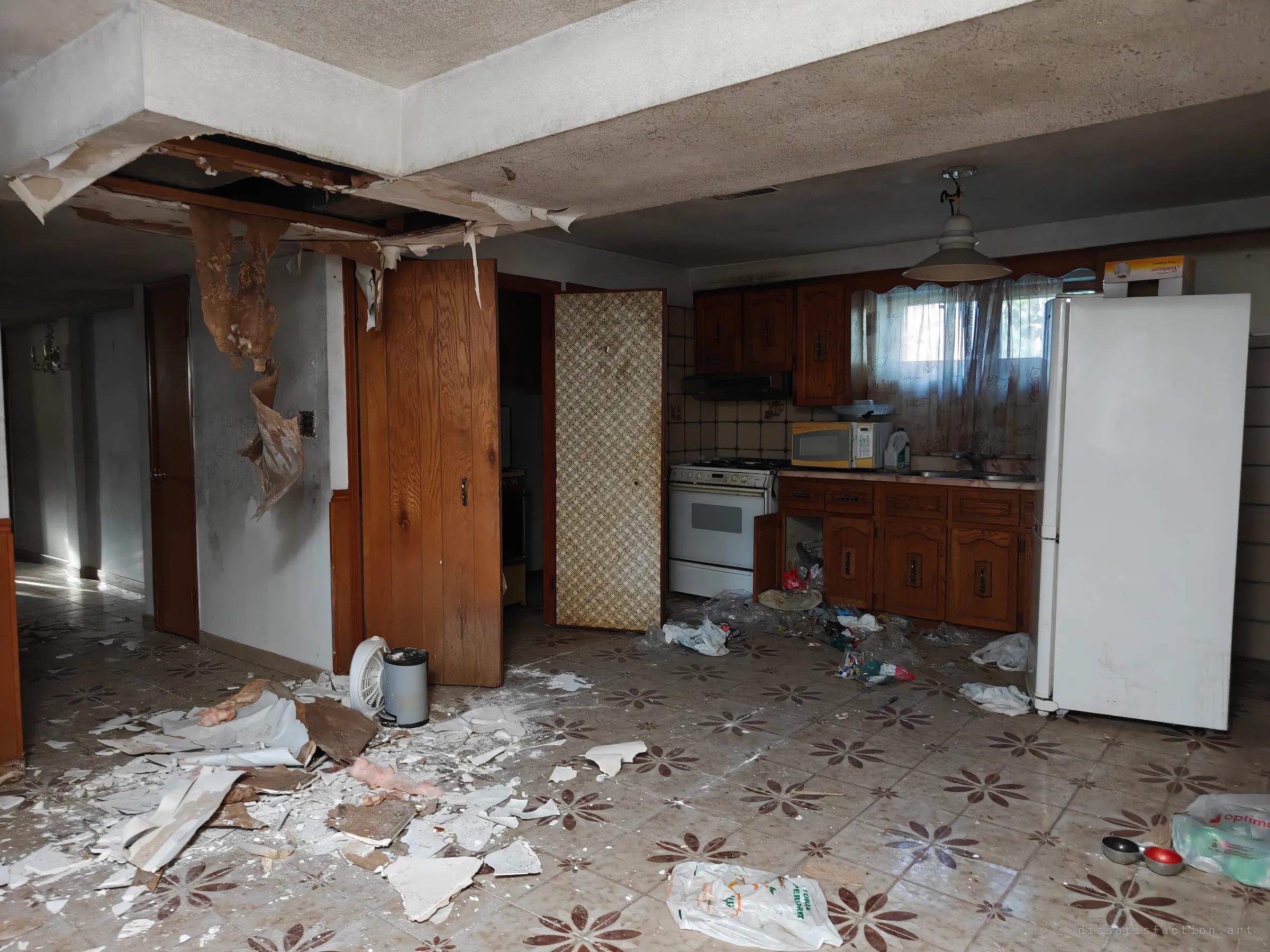 A collapsed ceiling hangs over a kitchen, with plaster and insulation scattered across a patterned tile floor and bags of rubbish strewn around a white fridge and wood cabinets.
