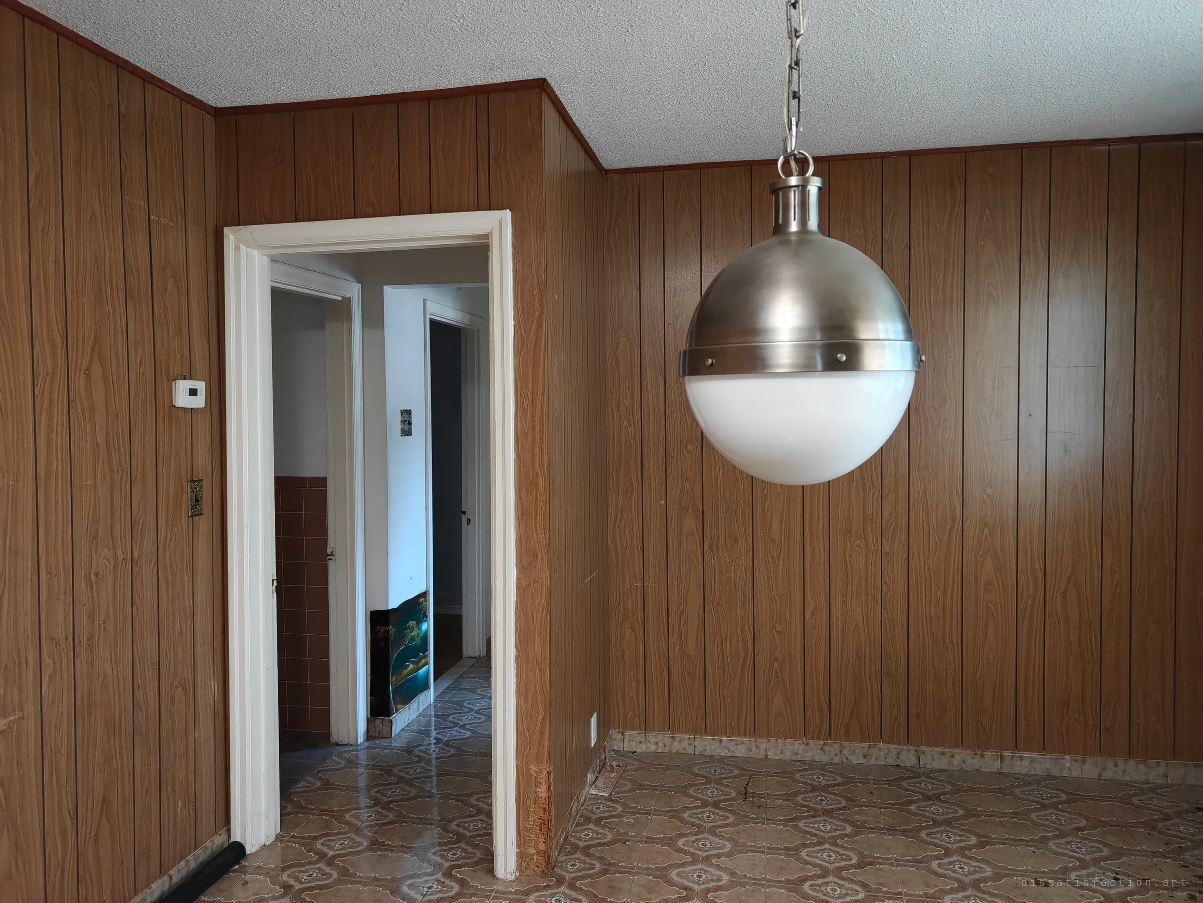 A wood-panelled dining room with a large brushed-steel and opal-glass globe pendant hanging beside an open doorway; patterned tile floor runs through to a hallway with a small painting on the floor.