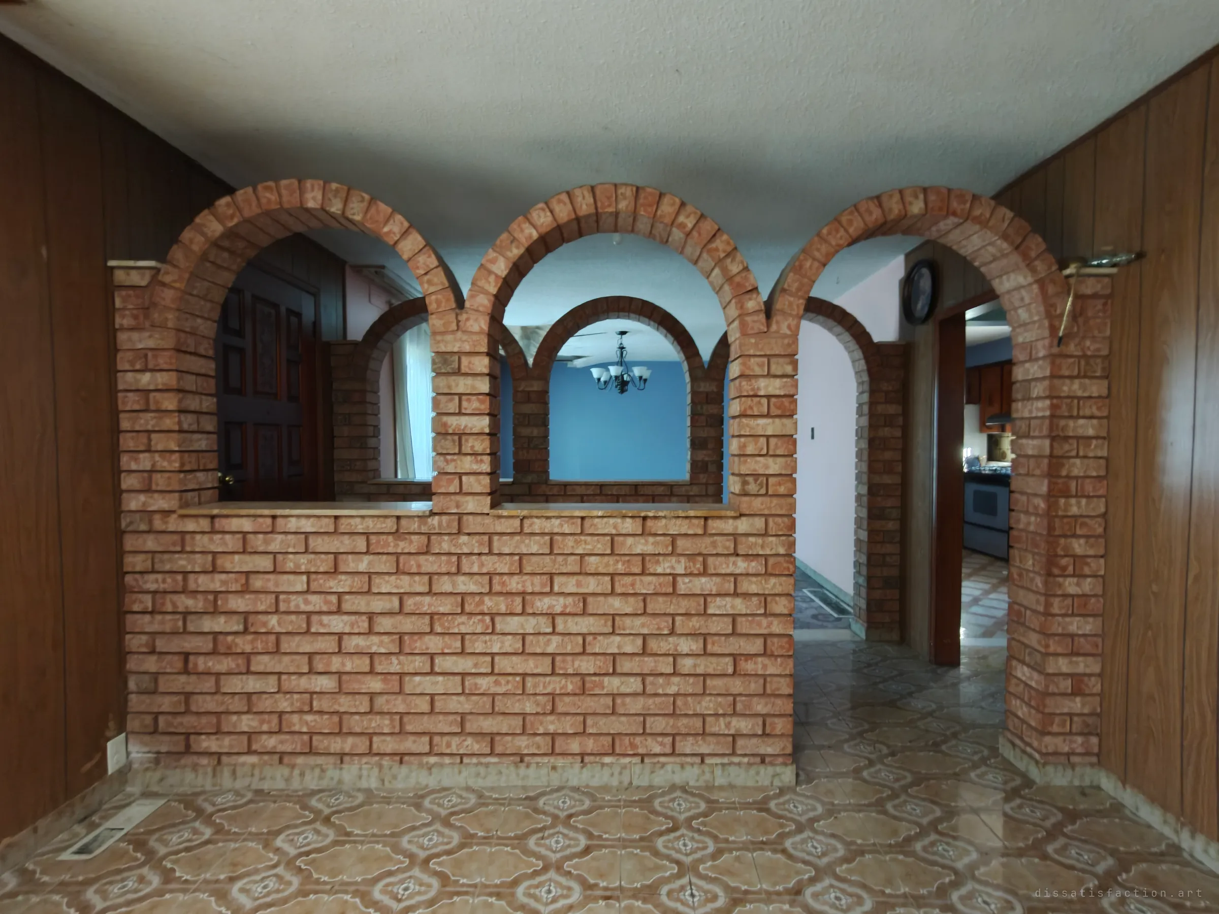 A brick partition wall with three arches and marble sills stands in a panelled room, with a chandelier, blue wall and kitchen visible through the openings, over patterned tile flooring.