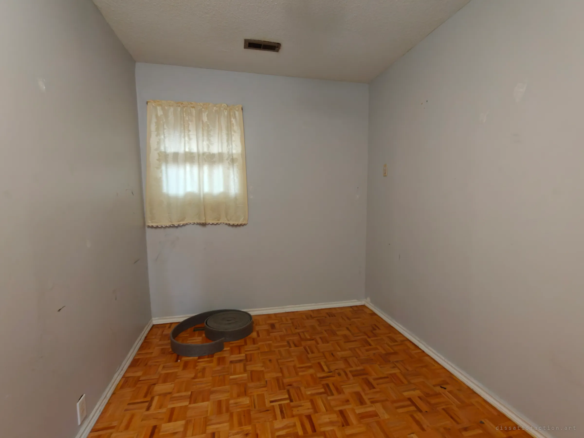 A small empty bedroom with grey-blue walls and a lace-curtained window. A coil of grey material lies on the parquet floor.