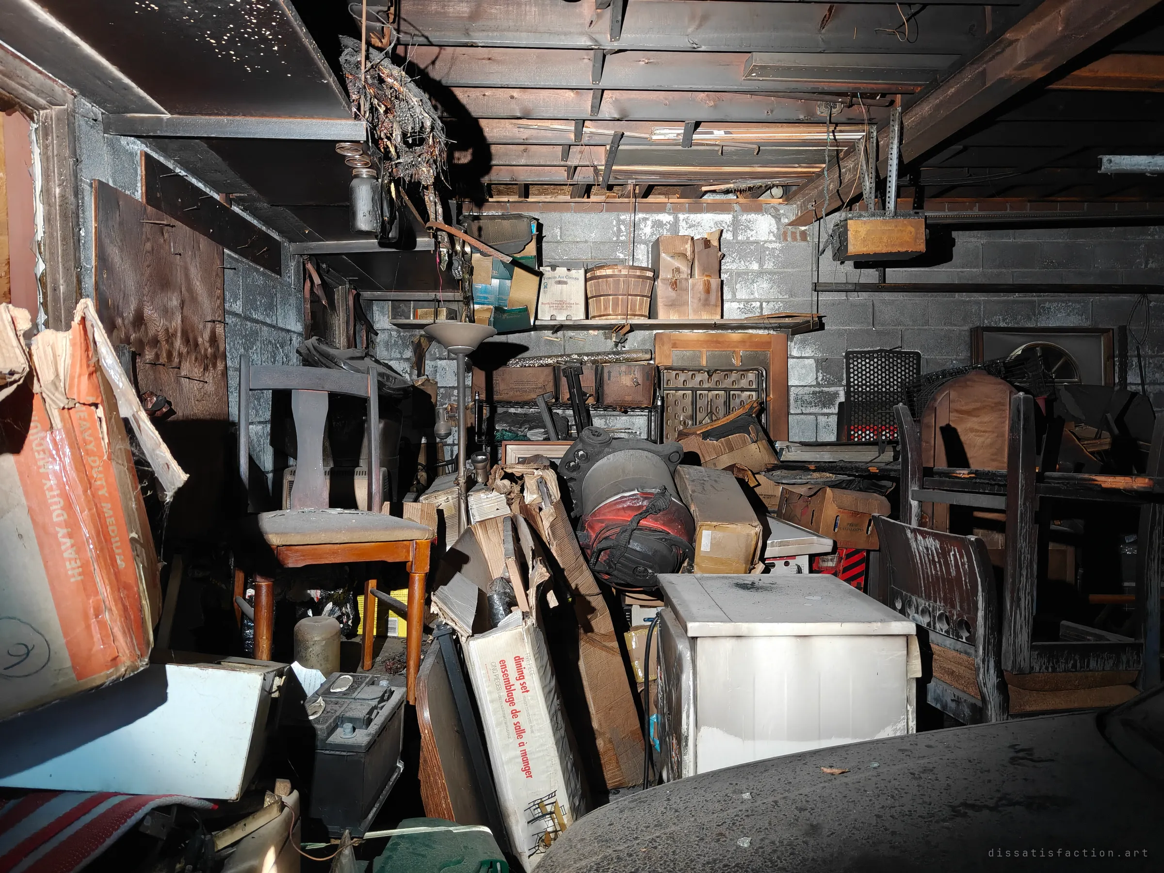 A cramped, soot-blackened garage interior packed with burnt chairs, cardboard boxes, a white appliance and a car battery under charred ceiling joists.