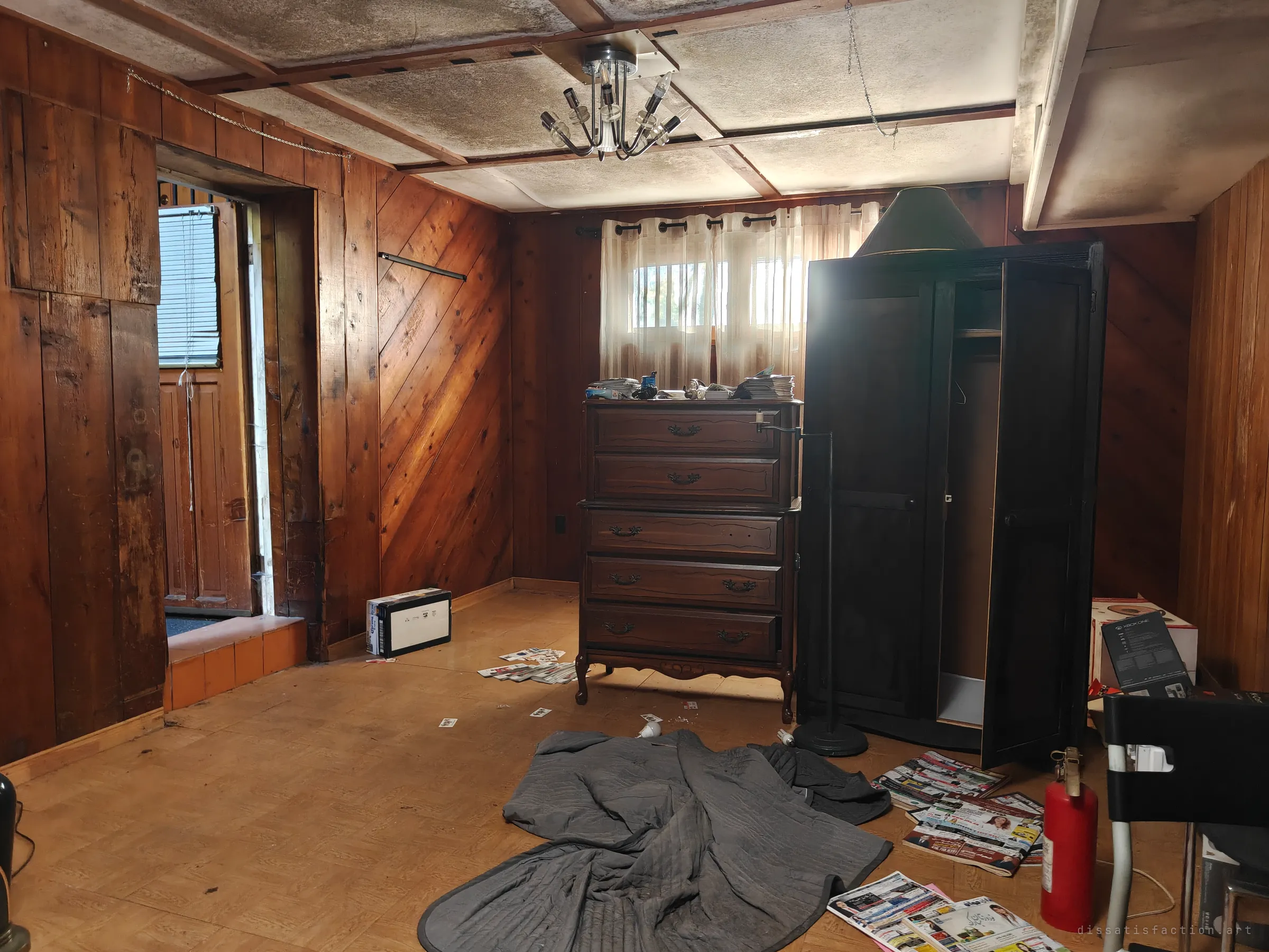 A wood-panelled room with a stained, sagging ceiling and a chandelier. A dark wardrobe and chest of drawers stand against the far wall, with a grey moving blanket, scattered flyers and a red fire extinguisher on the floor.