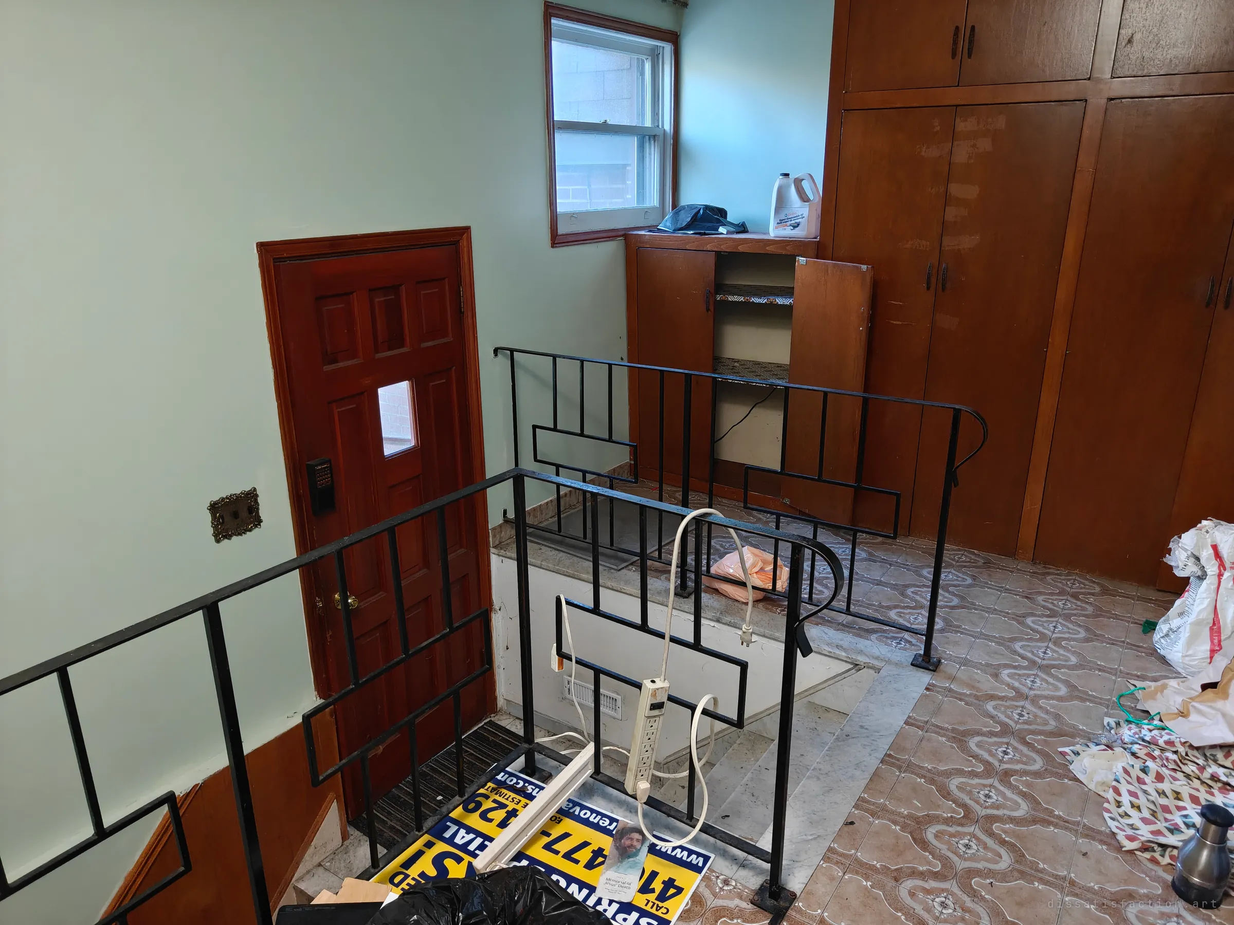 A landing at the top of a short flight of stairs, with a black wrought-iron railing, a dark red panelled door with a keypad lock, built-in wooden cupboards, and a fallen renovation sign, power strip and rubbish bags left on the floor.