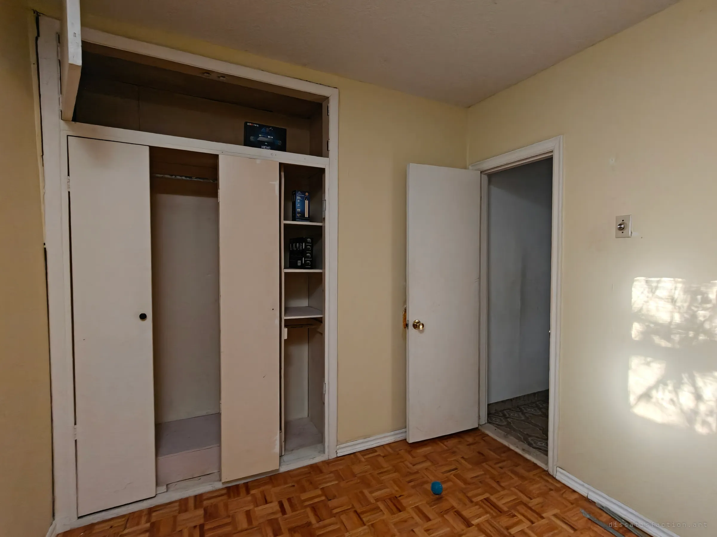 A yellow bedroom with a built-in closet, its doors pulled aside to show a few leftover product boxes on the shelves. An open door leads to a hallway, and a small blue ball lies on the parquet floor.