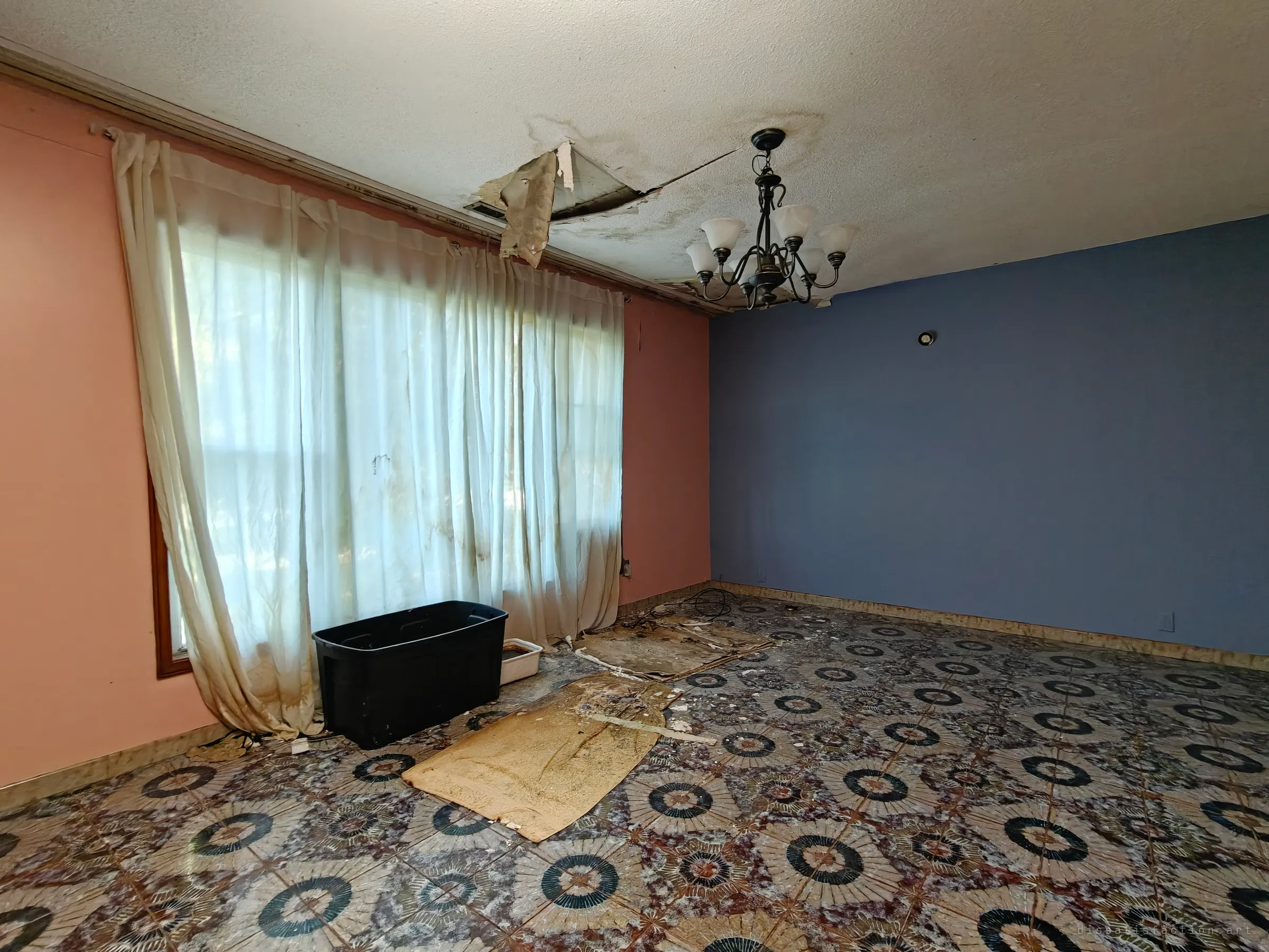 A gutted living room with a pink wall, a blue wall and long stained curtains over a window. A ceiling panel has collapsed, a chandelier still hangs, and the patterned floor is covered in debris and a black storage tub.