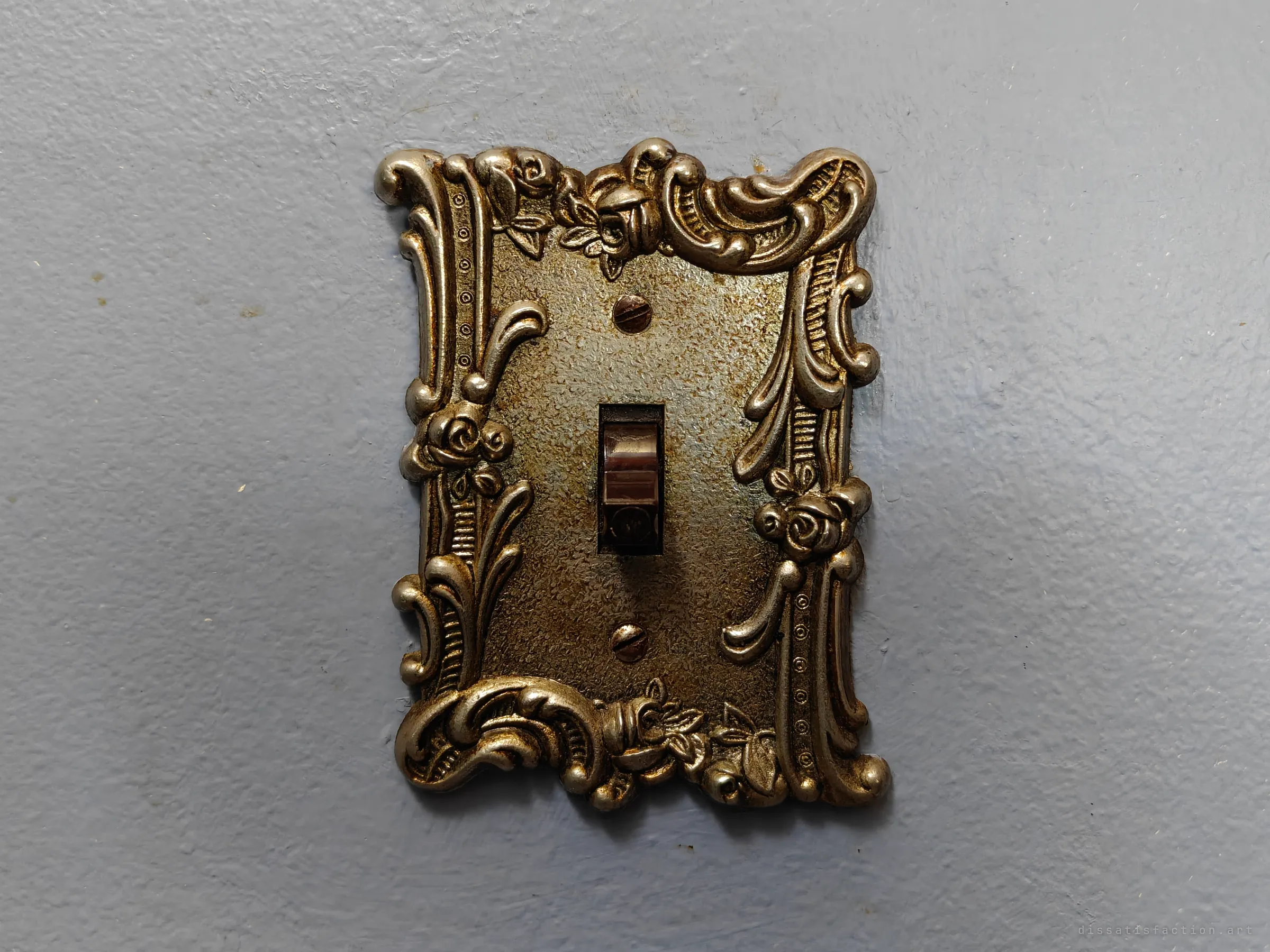 A close view of an ornate tarnished brass light-switch plate with rose and scroll detail on a pale blue painted wall.