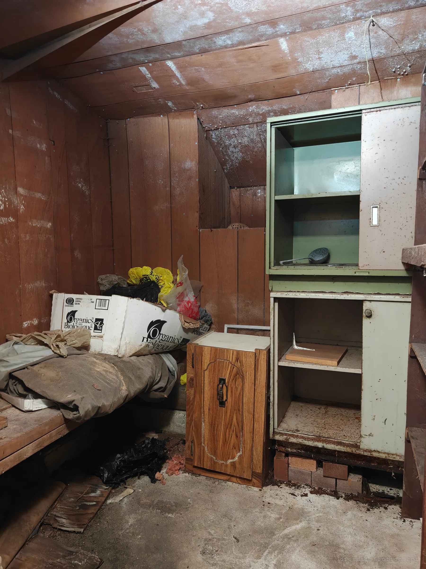 A small wood-panelled storage room with a mould-streaked sloped ceiling. A green metal cabinet, a wooden side cupboard and a box of discarded bags and blankets sit against the walls.