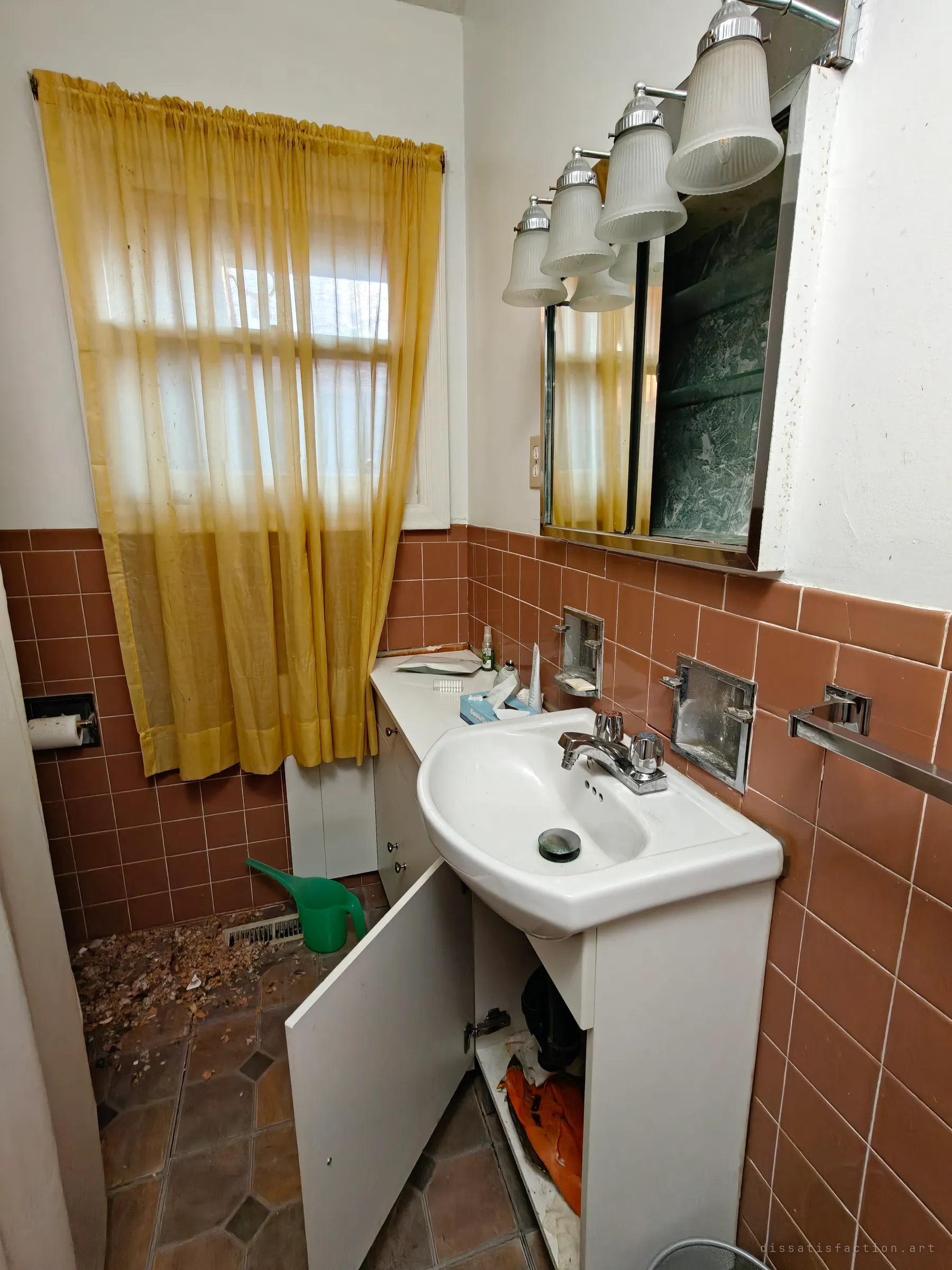 A small bathroom in brown tile and a yellowed curtain, its vanity door hung open over a floor scattered with debris and a green watering can.