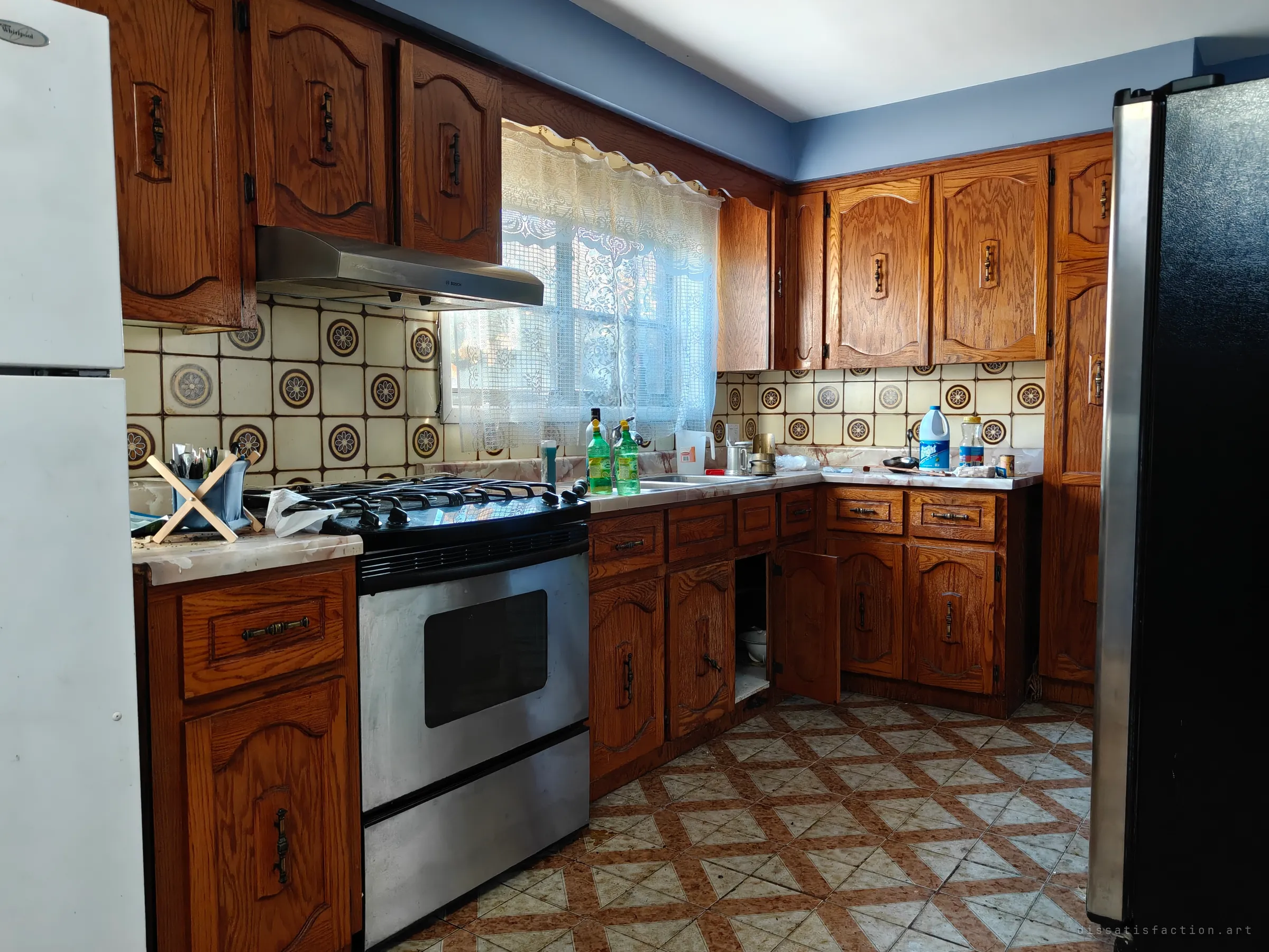 A dated kitchen of stained oak cabinets, a gas range and cream medallion backsplash tiles, with a lace-curtained window, bottles and clutter left on the counter and one cupboard standing open.