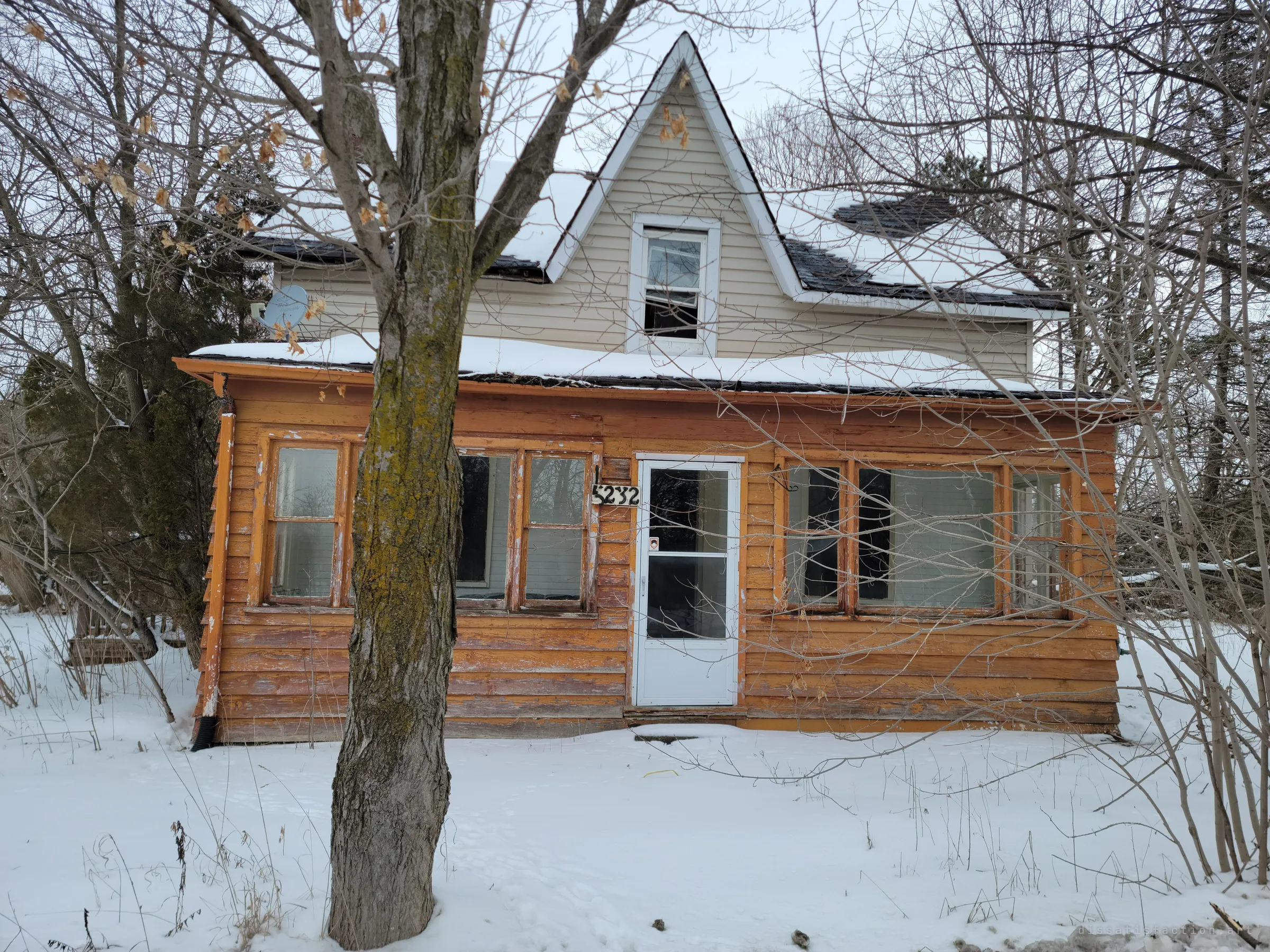The front of a small abandoned house in snow, its weathered orange board porch and white storm door half hidden behind a bare tree. Snow on the roof and a gaping upper window show how long it has sat empty.