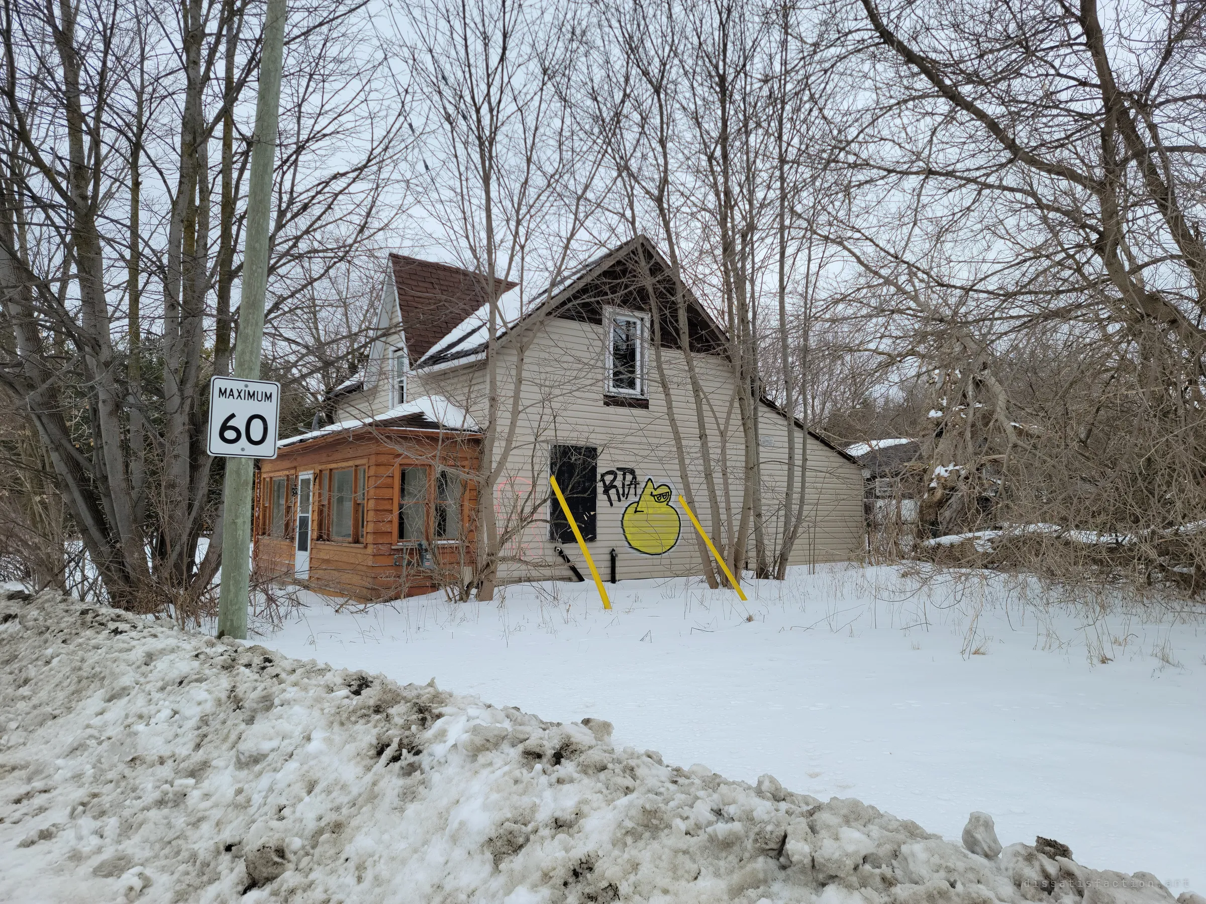 The house seen from the plowed roadside, a boarded window on its pale siding tagged with a yellow cartoon face and propped by yellow poles. It sits back among bare trees behind a snowbank.