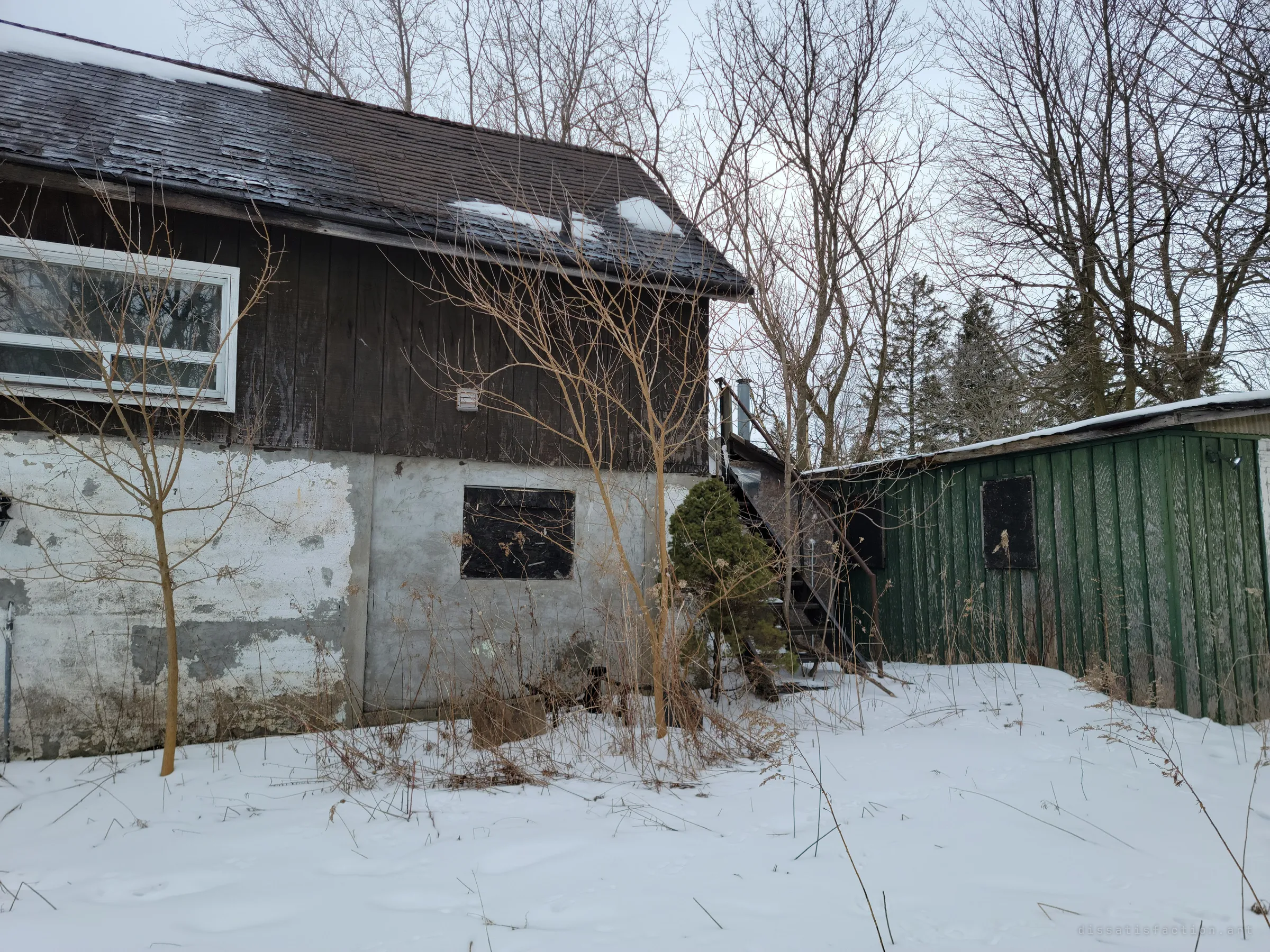 Closer to the outbuilding: peeling shingles, a boarded window in flaking white block and a rusted exterior staircase beside a green board shed. Weeds poke through the snow along the wall.