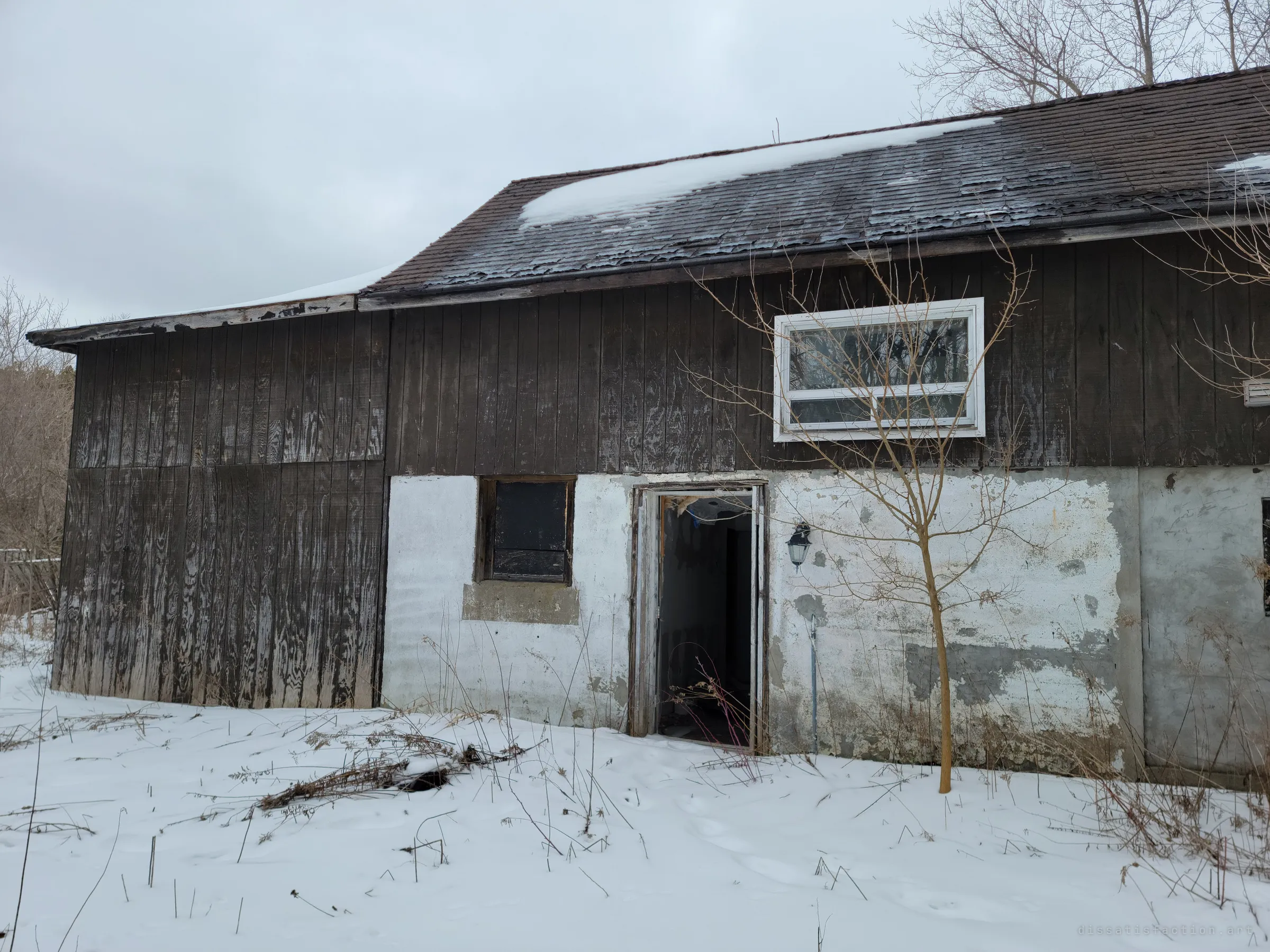 The long side of the outbuilding, its door hanging open onto a dark interior under a patchy, curling shingle roof. A lone sapling and a porch lamp stand against the stained white wall.