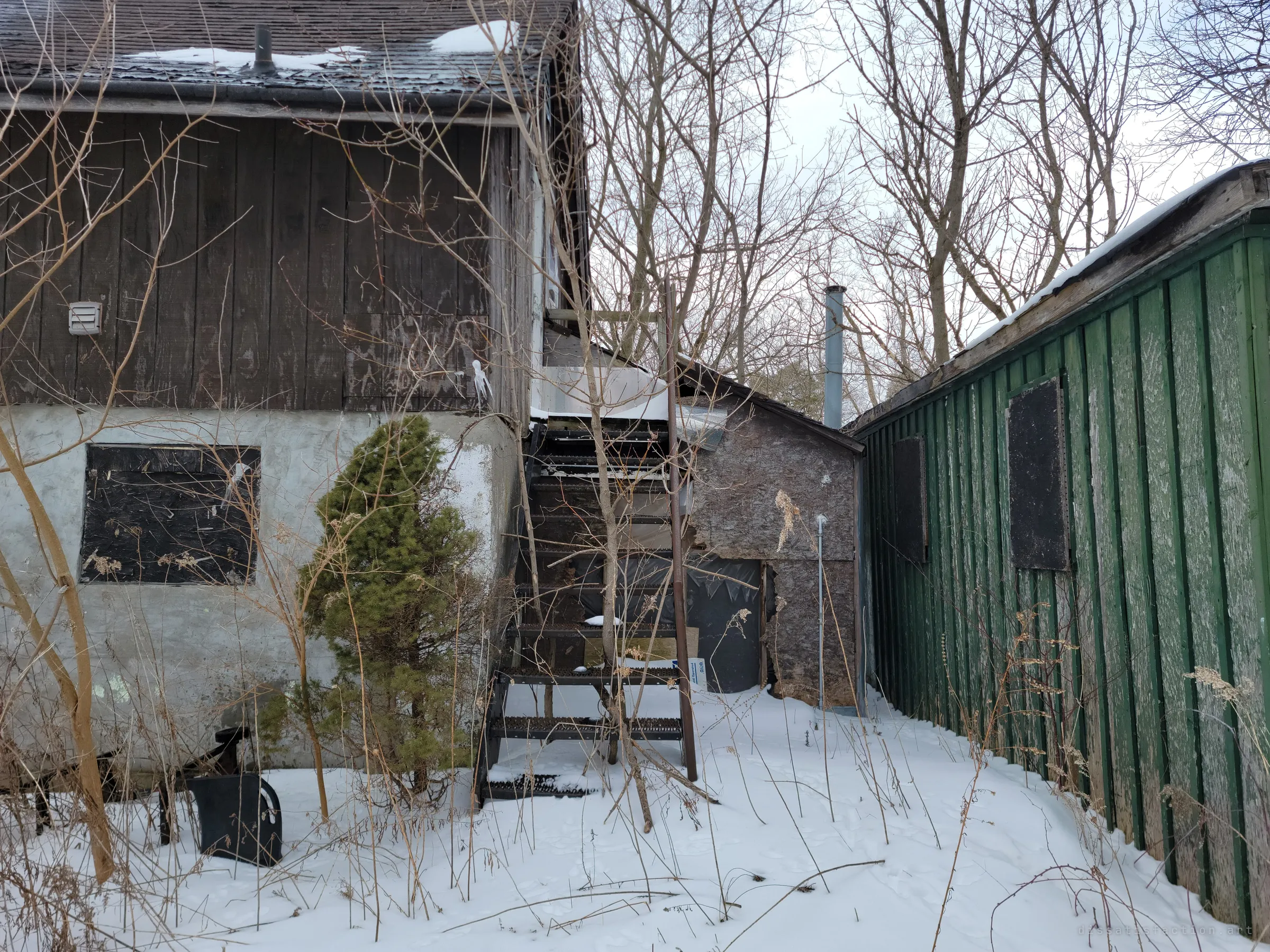 A narrow gap between the outbuilding and the green shed where a steel staircase climbs to an upper door. Boarded windows, a small evergreen and drifted snow fill the passage.