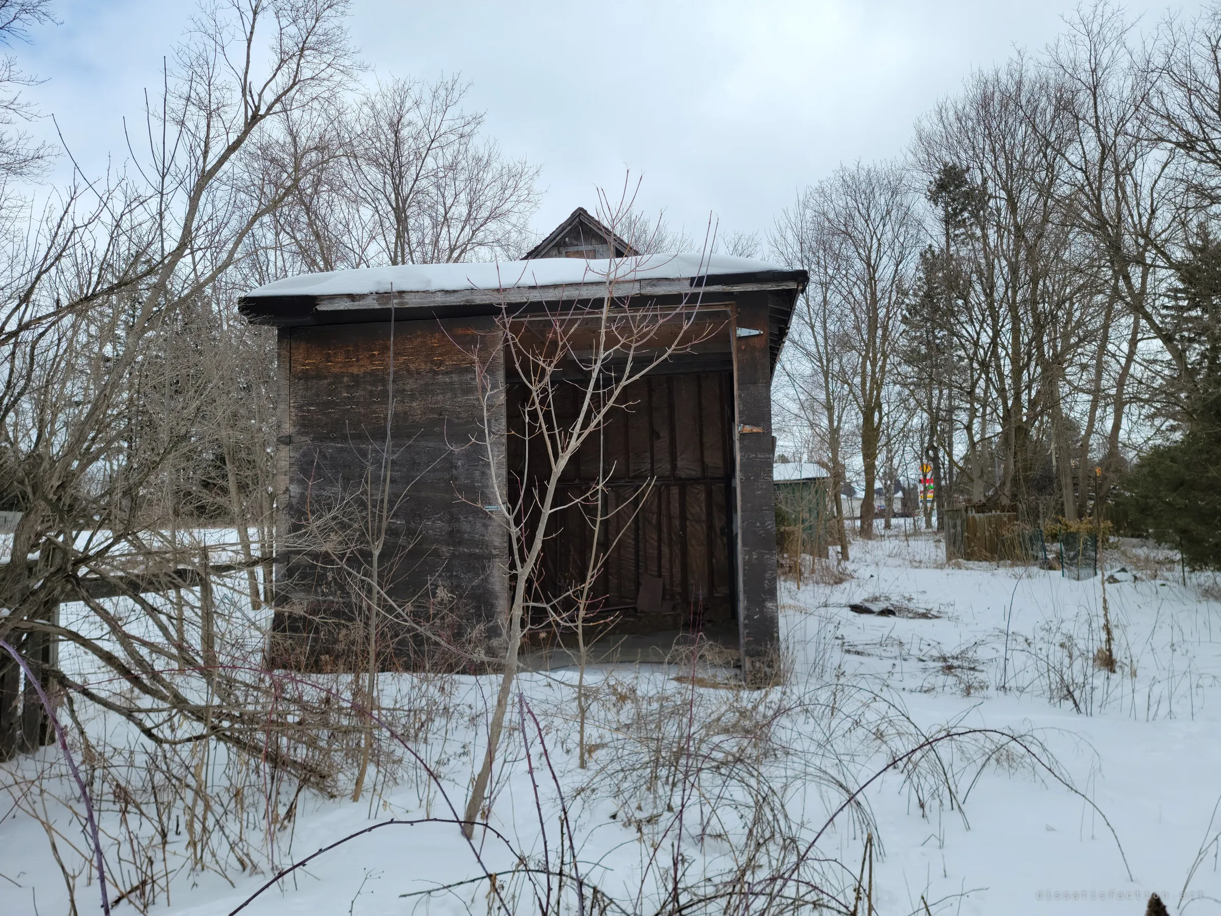 An open-fronted wooden shed with a small cupola, snow heaped on its flat roof and its empty bay facing a tangle of bare brush. It stands at the edge of the lot among old trees.