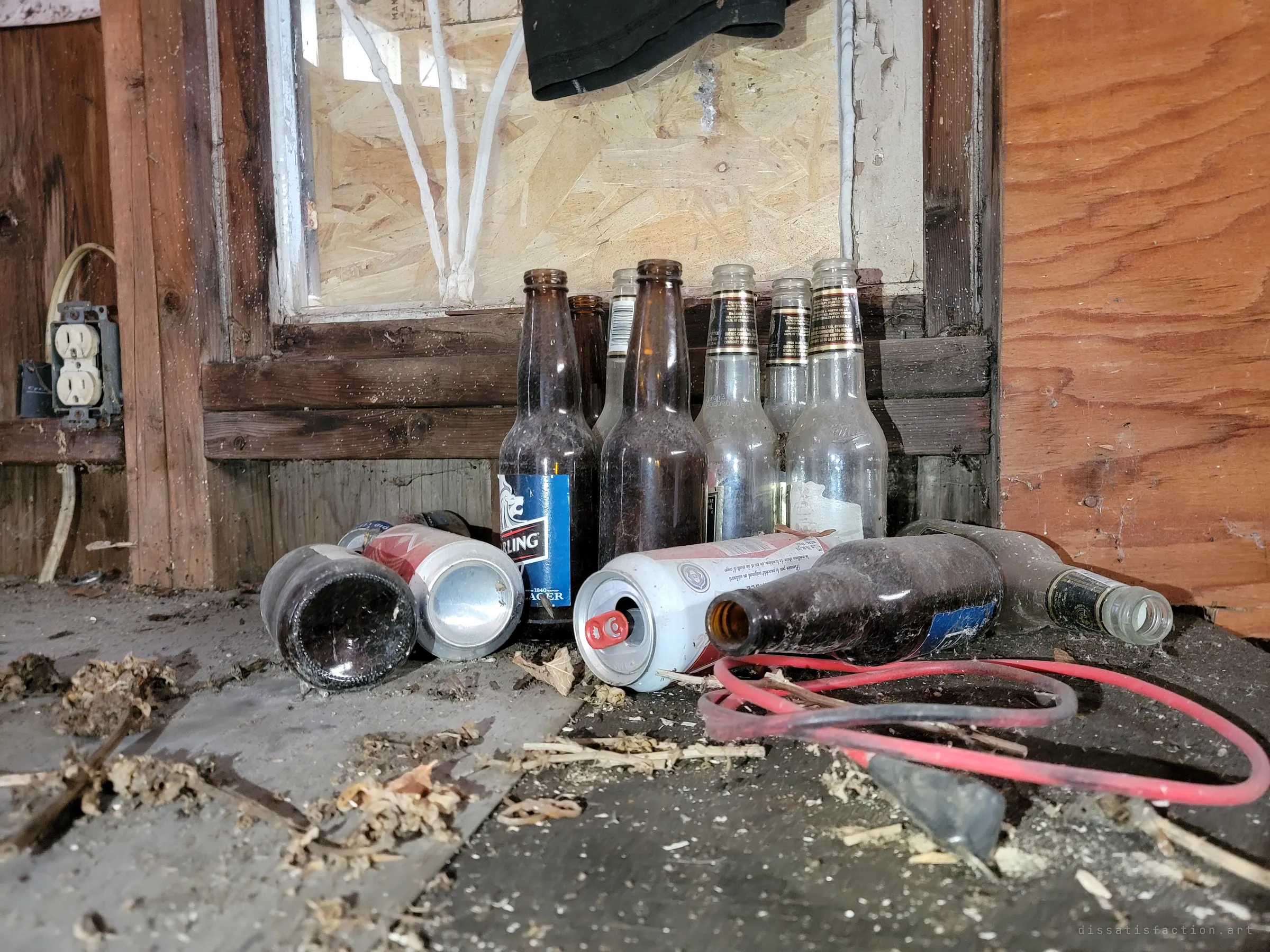 Dusty empty beer bottles and crushed cans lined up under a boarded window, beside a red jumper cable and an old wall outlet. Someone drank here long after the owners left.