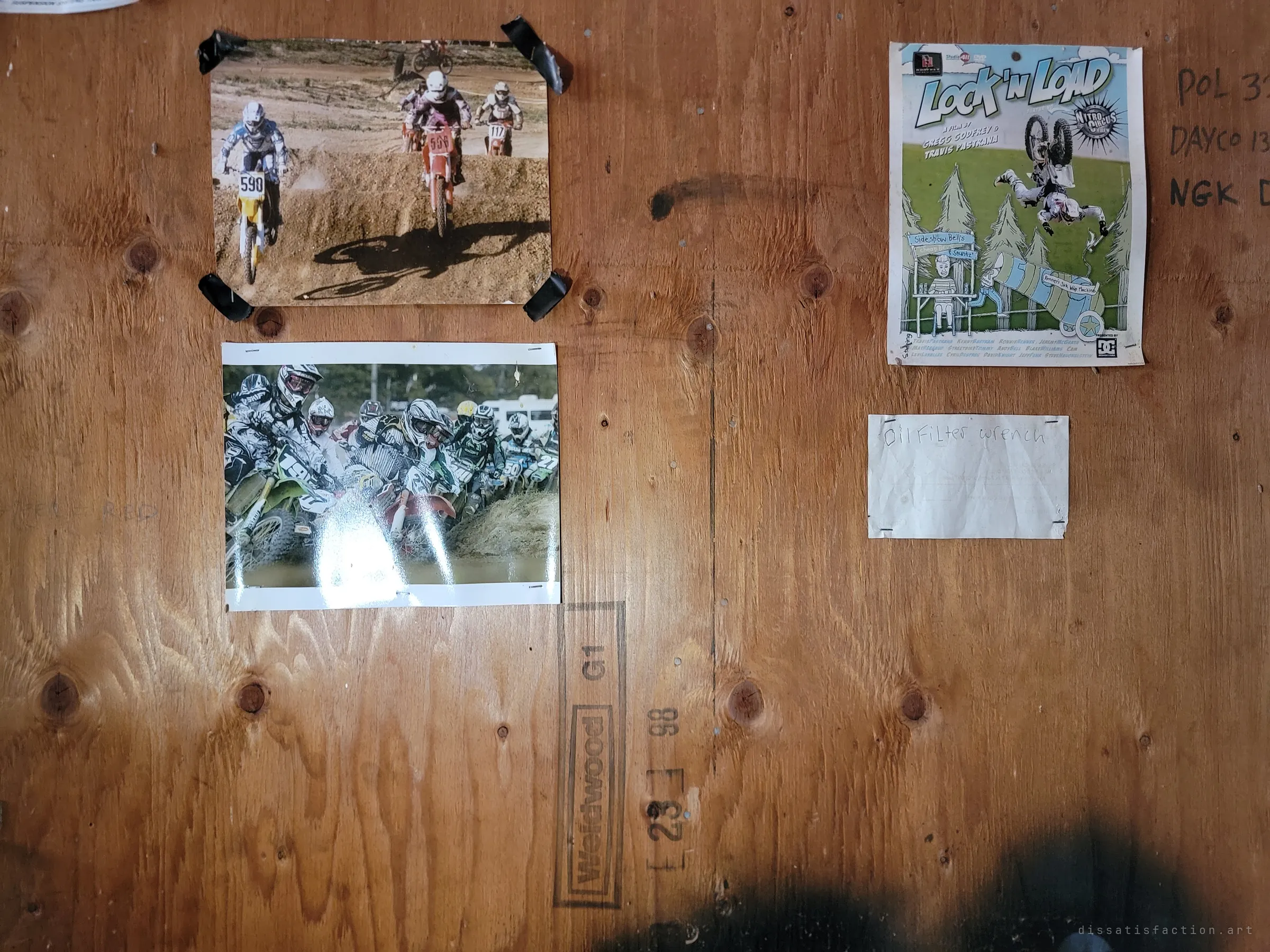 A plywood garage wall with taped-up motocross photos, a stunt-film poster and a handwritten note for an oil filter wrench, plus parts numbers scrawled in marker. Traces of whoever once worked on bikes here.