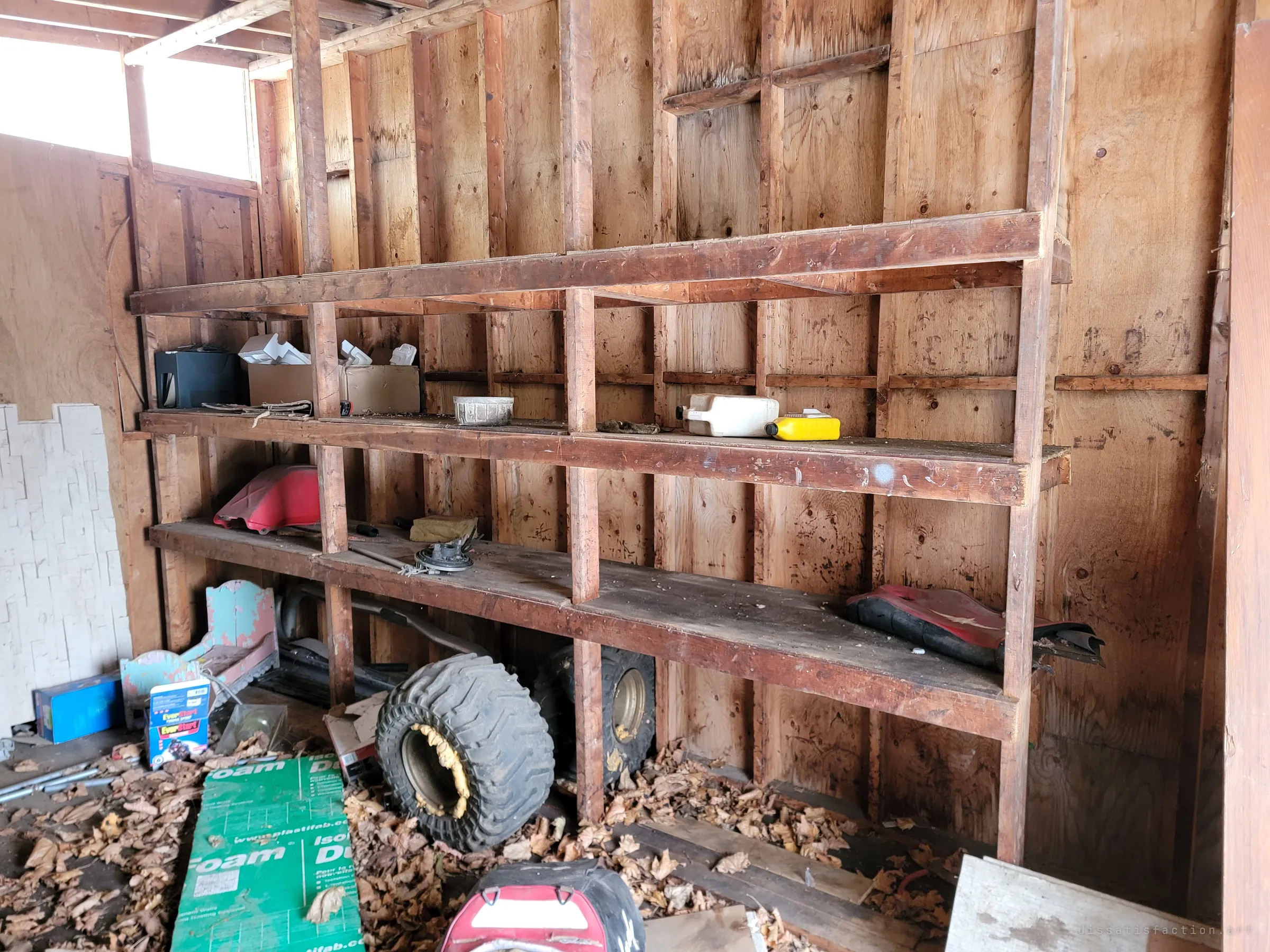 Rough timber shelving along a plywood garage wall, holding old boxes, jugs and a dirt-bike tank, with loose wheels, foam board and dead leaves piled on the floor below. The leftovers show the place was used as a working garage before it was abandoned.