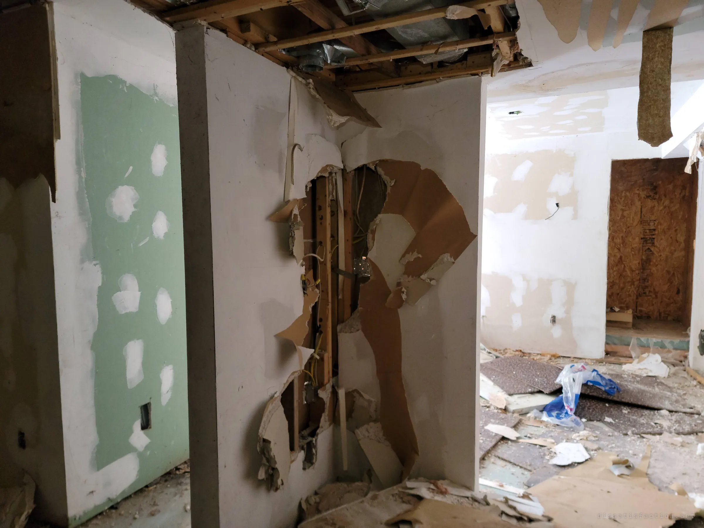 A drywall column ripped open to its wiring and studs, between a green-board wall dotted with patch marks and a debris-strewn room with a boarded-up doorway. The half-finished patching and torn walls show a renovation that was abandoned partway.