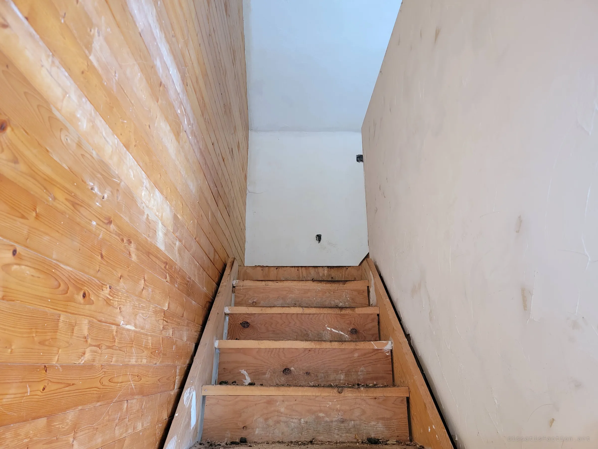 Looking up a narrow, steep plywood staircase between a knotty pine wall and a bare white wall, toward an empty landing. The cramped stair suggests a converted attic floor above.