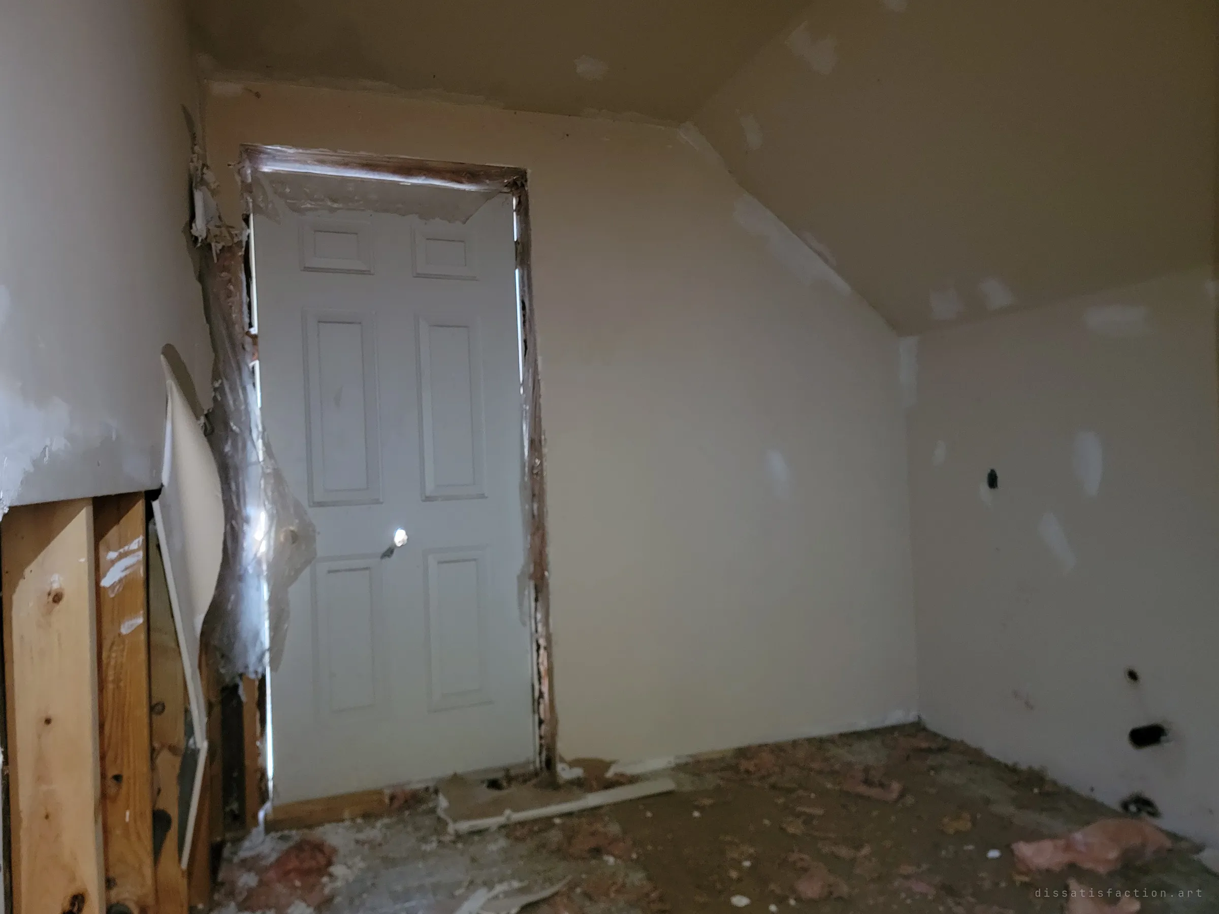 A dim attic room under a sloped ceiling, with a panel door wrapped in torn plastic sheeting and scattered insulation on the floor. Light leaking around the door frame is the only thing lighting the room.