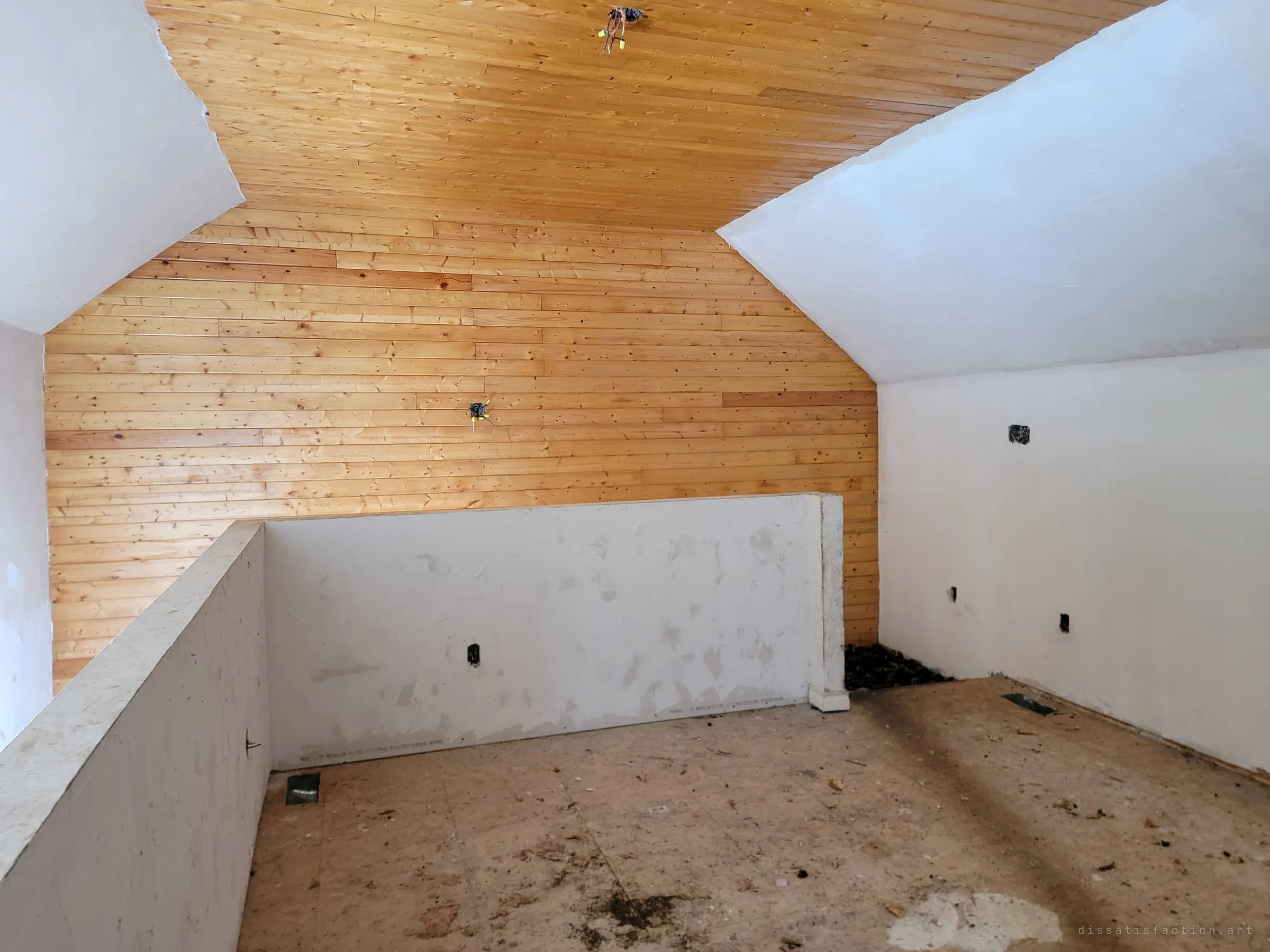 An empty upstairs room under a pine-clad gable, with a low drywall half-wall around the stair opening and bare subfloor. Dangling wires where the lights used to be show it was stripped before it was finished.
