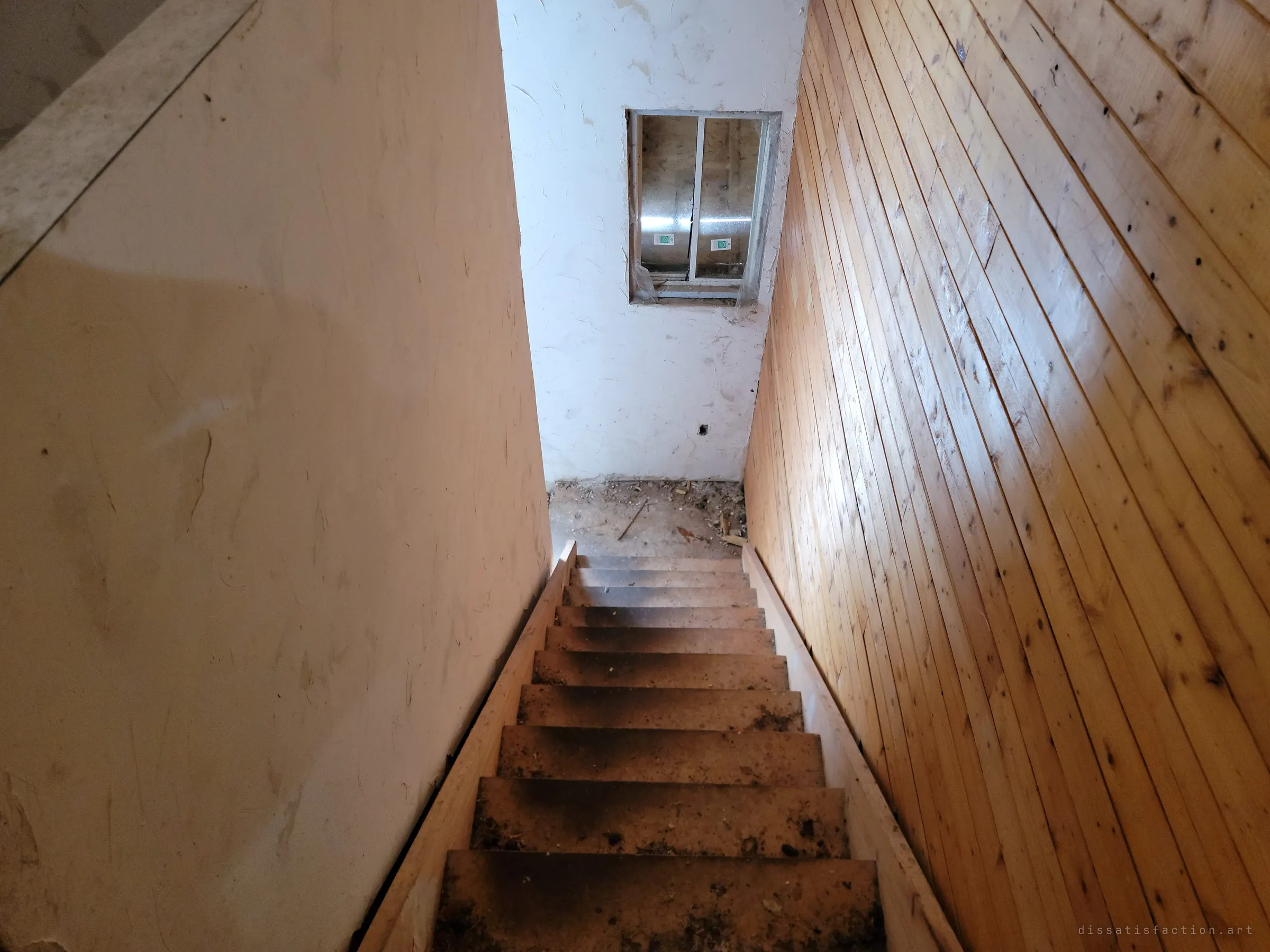 Looking down a steep, dirty wooden staircase between a pale wall and pine panelling, toward a landing with a still-stickered new window set into the wall. The unused window hints at a renovation that stopped suddenly.