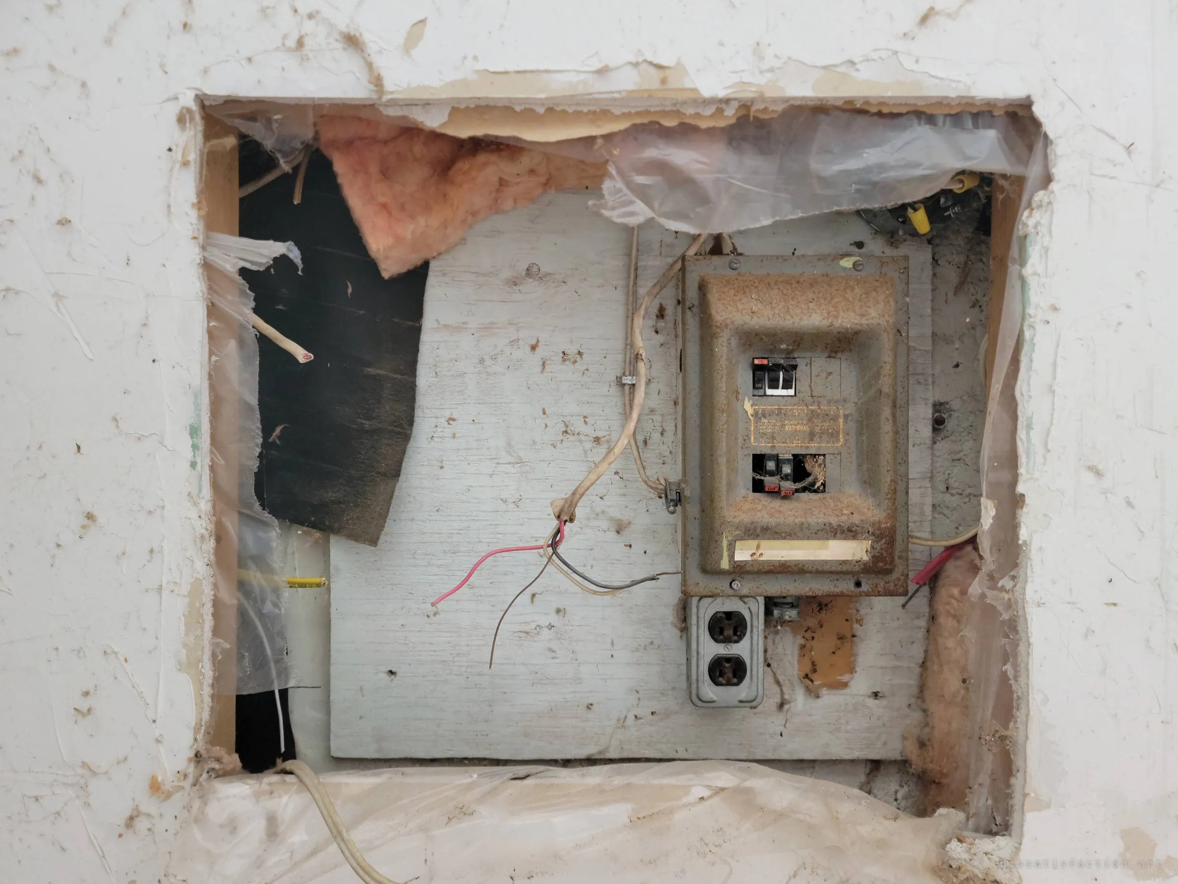 A rusted breaker panel and a bare outlet sit exposed behind a hole hacked through the drywall, cut wires and pink insulation spilling out around them. The house has been gutted down to its wiring.