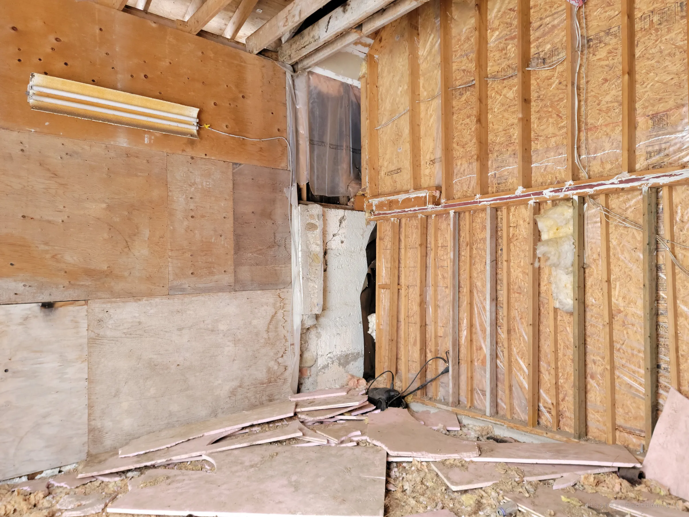 A corner stripped to the studs: patched plywood on one side, bare OSB and torn vapour barrier on the other, with broken pink foam board piled on the floor. Shows how far the renovation or stripping got before it stopped.
