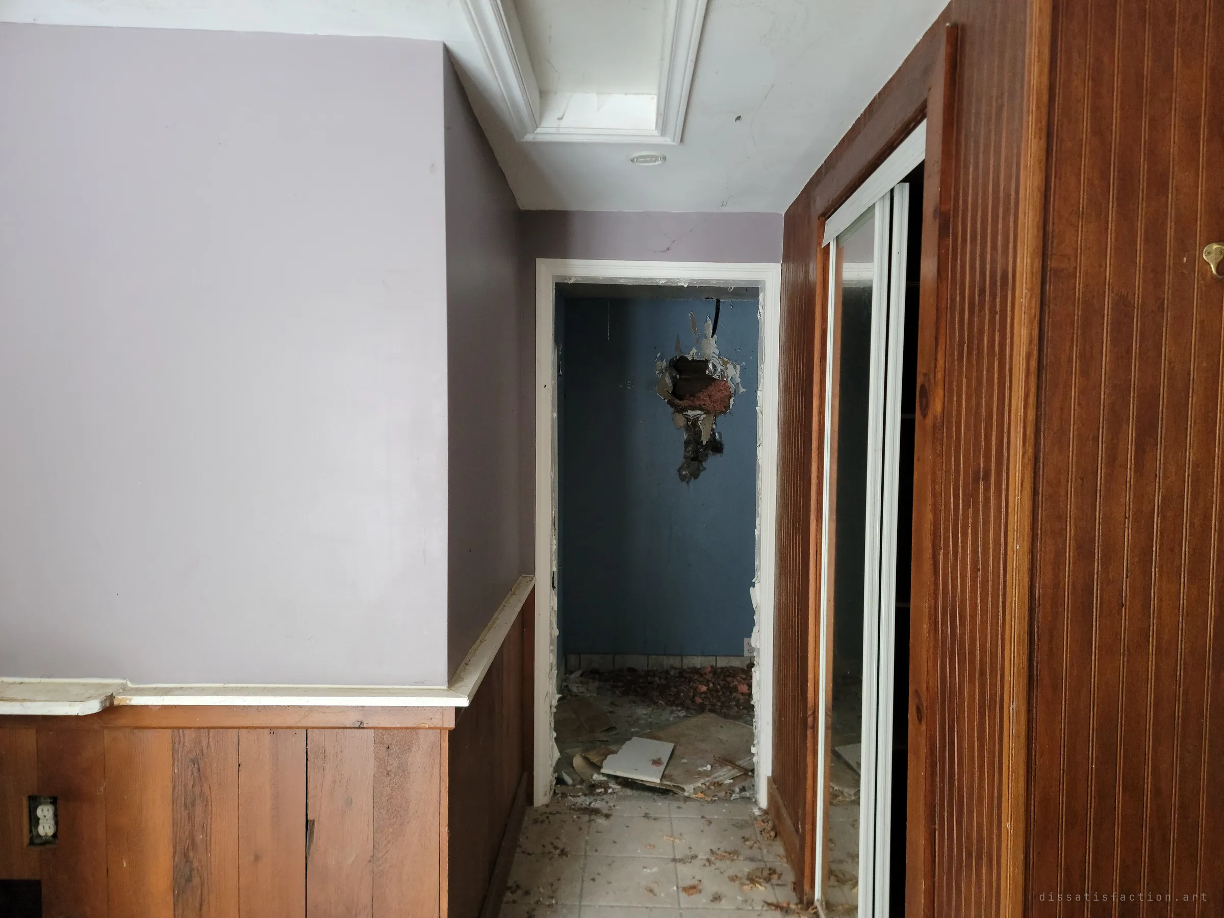 A lavender hallway with wood wainscoting leads to a small blue room with a hole punched through its wall and debris on the floor. A mirrored closet door stands open on the right.