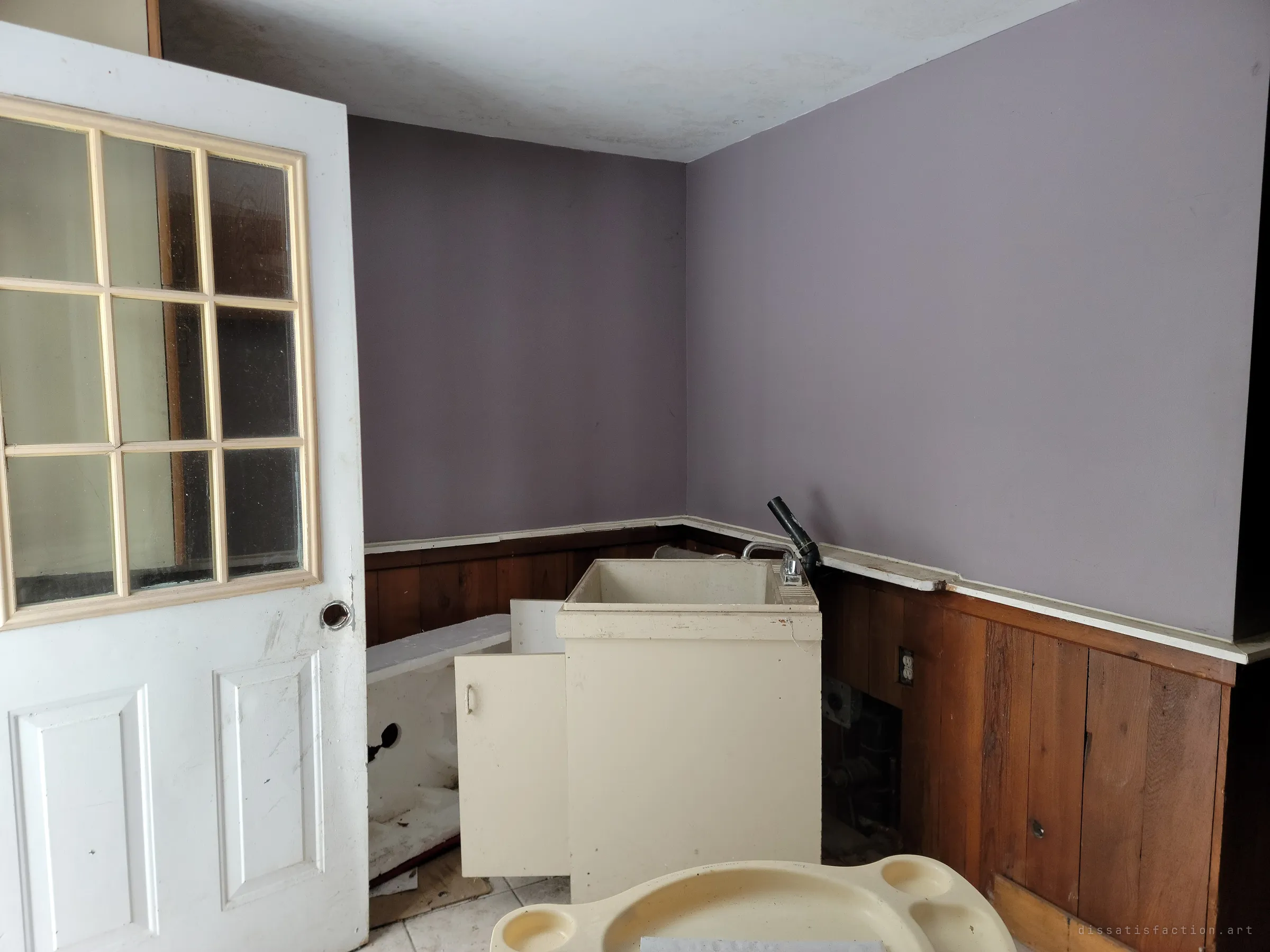 A lavender room with a half-glazed door swung open, a freestanding laundry sink pulled from the wall, and a plastic tray in the foreground. Pipes are exposed where fixtures were ripped out.