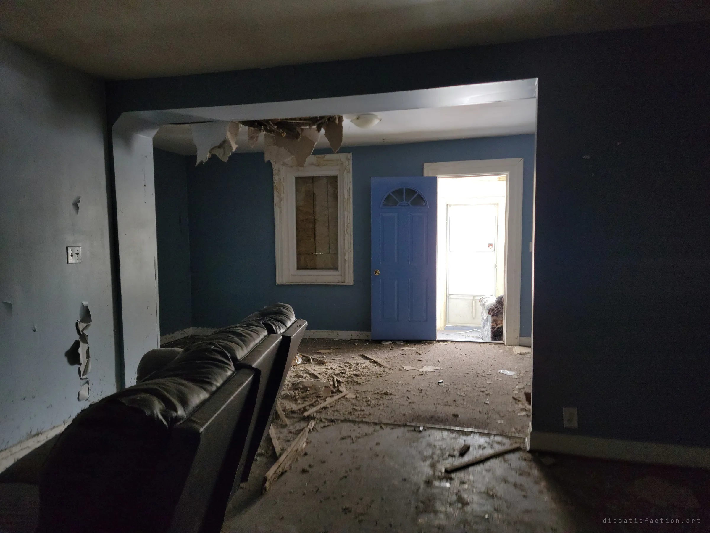 A dark living room with a leather sofa facing a collapsed ceiling, a boarded window and a blue door opening onto a bright enclosed porch. Light from the porch is the only thing left in the room.
