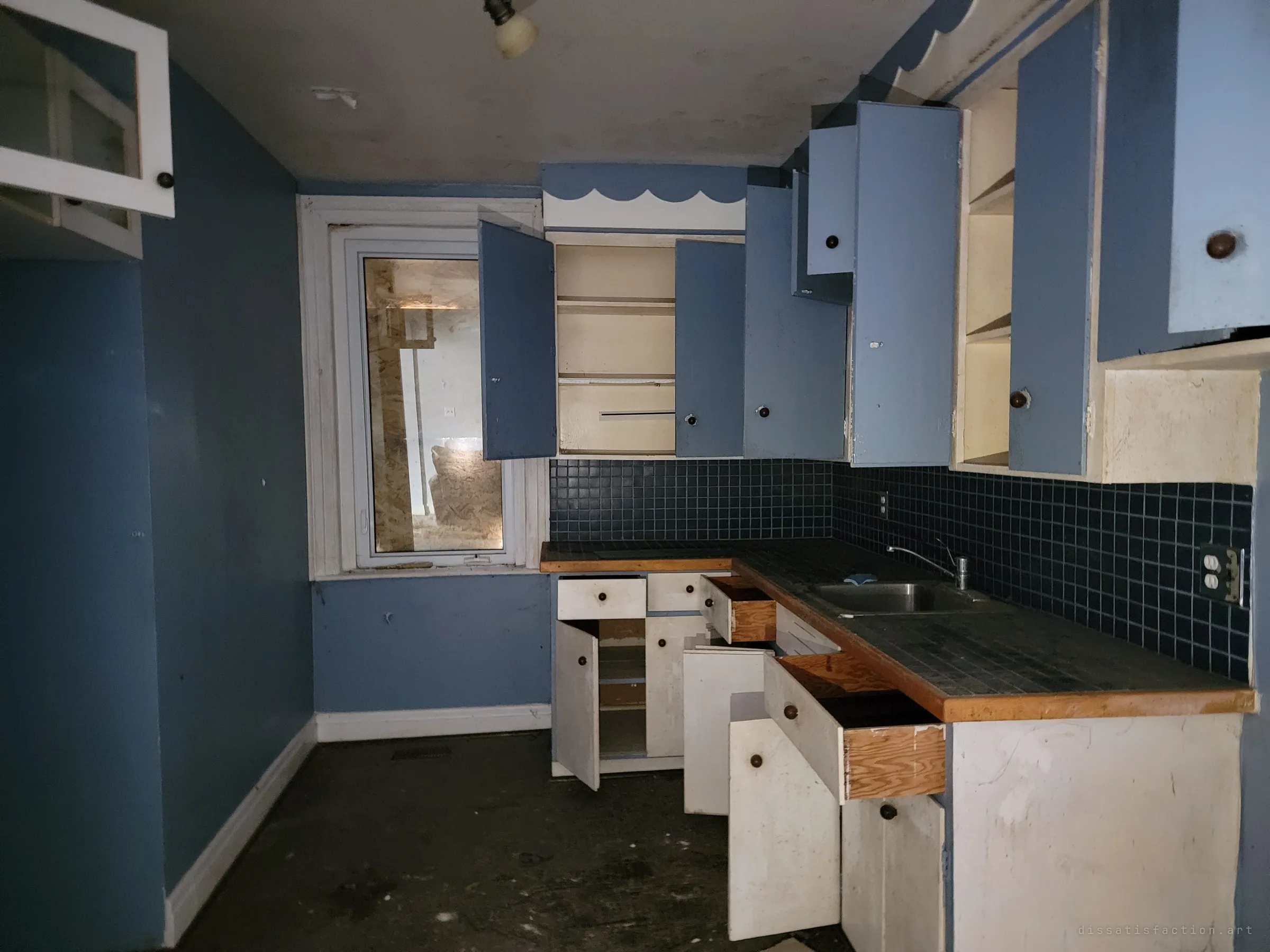 A blue kitchen with every cabinet and drawer hanging open, a dark tiled counter and a boarded window beyond. It looks like the house was searched or scavenged.