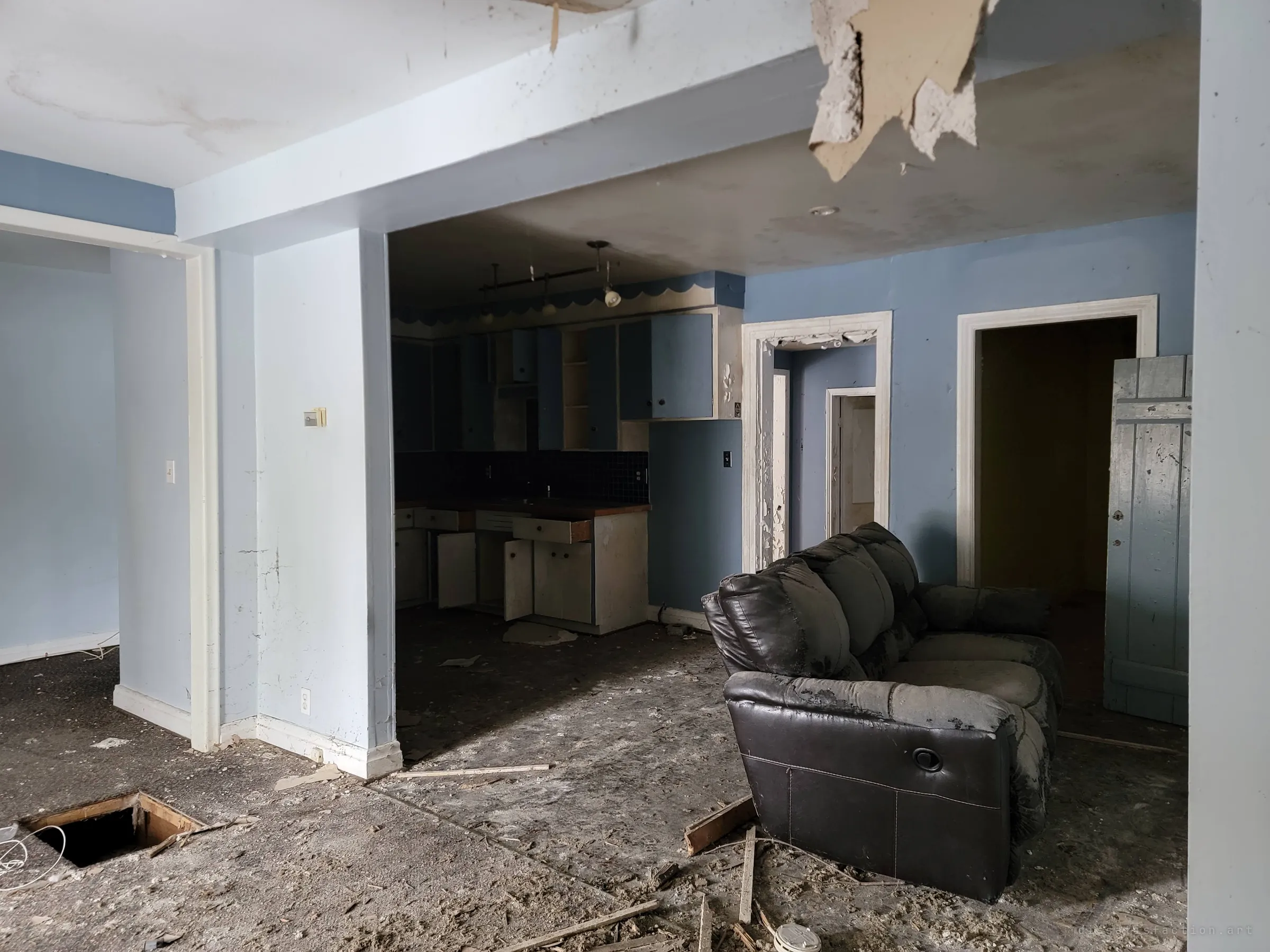 An open living area with a torn ceiling panel, a hole cut in the floor and a dusty leather recliner, the stripped blue kitchen visible through the opening. Several doorways lead further into the house.