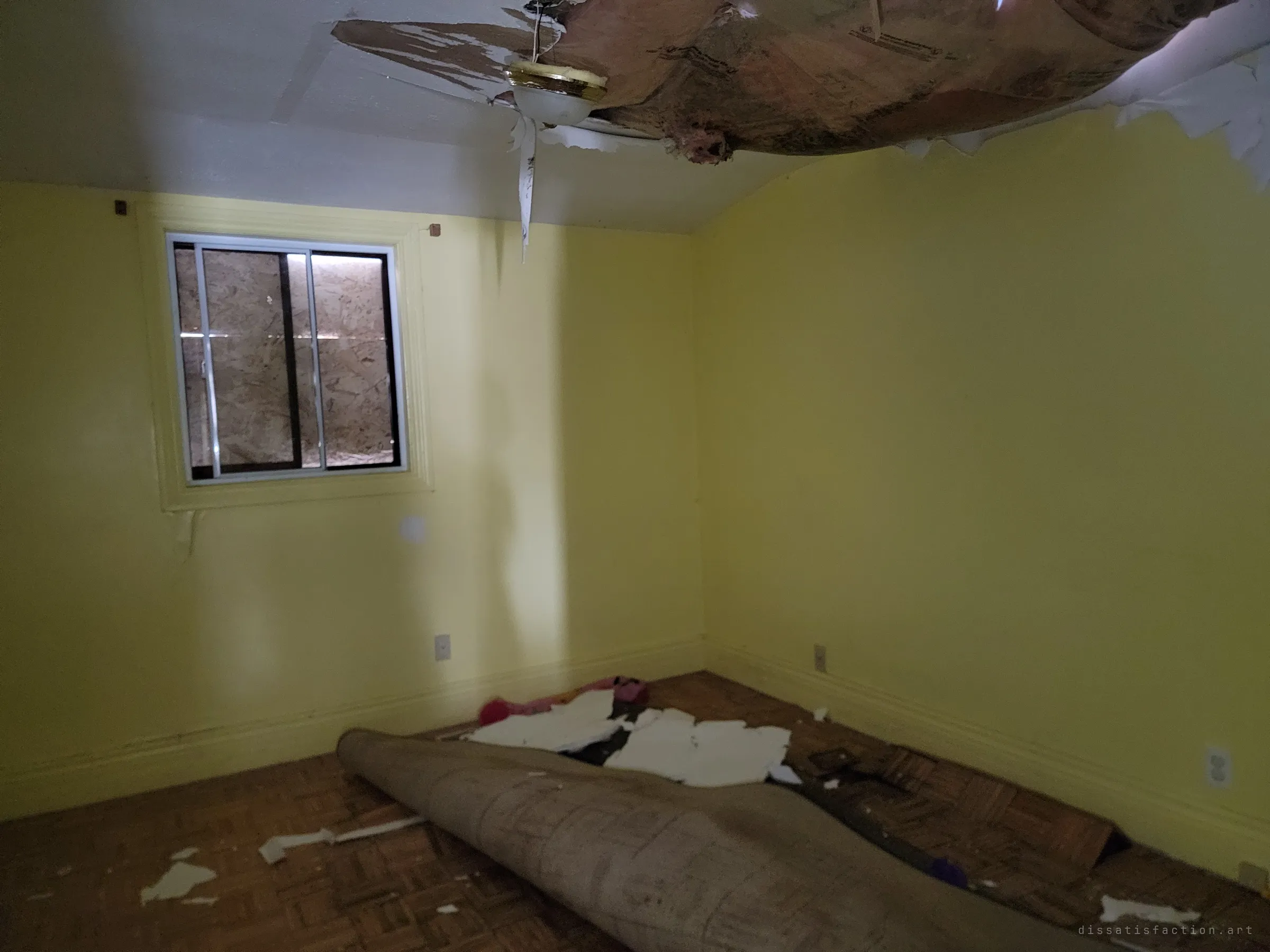 A yellow bedroom with the ceiling sagging open around a hanging light fixture and a rolled-up carpet on the parquet floor. The window is boarded from outside.