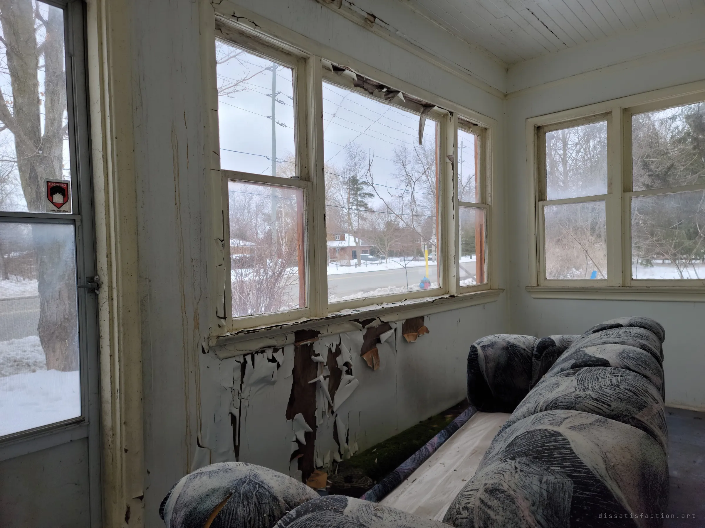 An enclosed winter porch with peeling paint below the windows and a patterned couch left behind, snow outside. The sunroom still feels lived-in while the walls come apart.